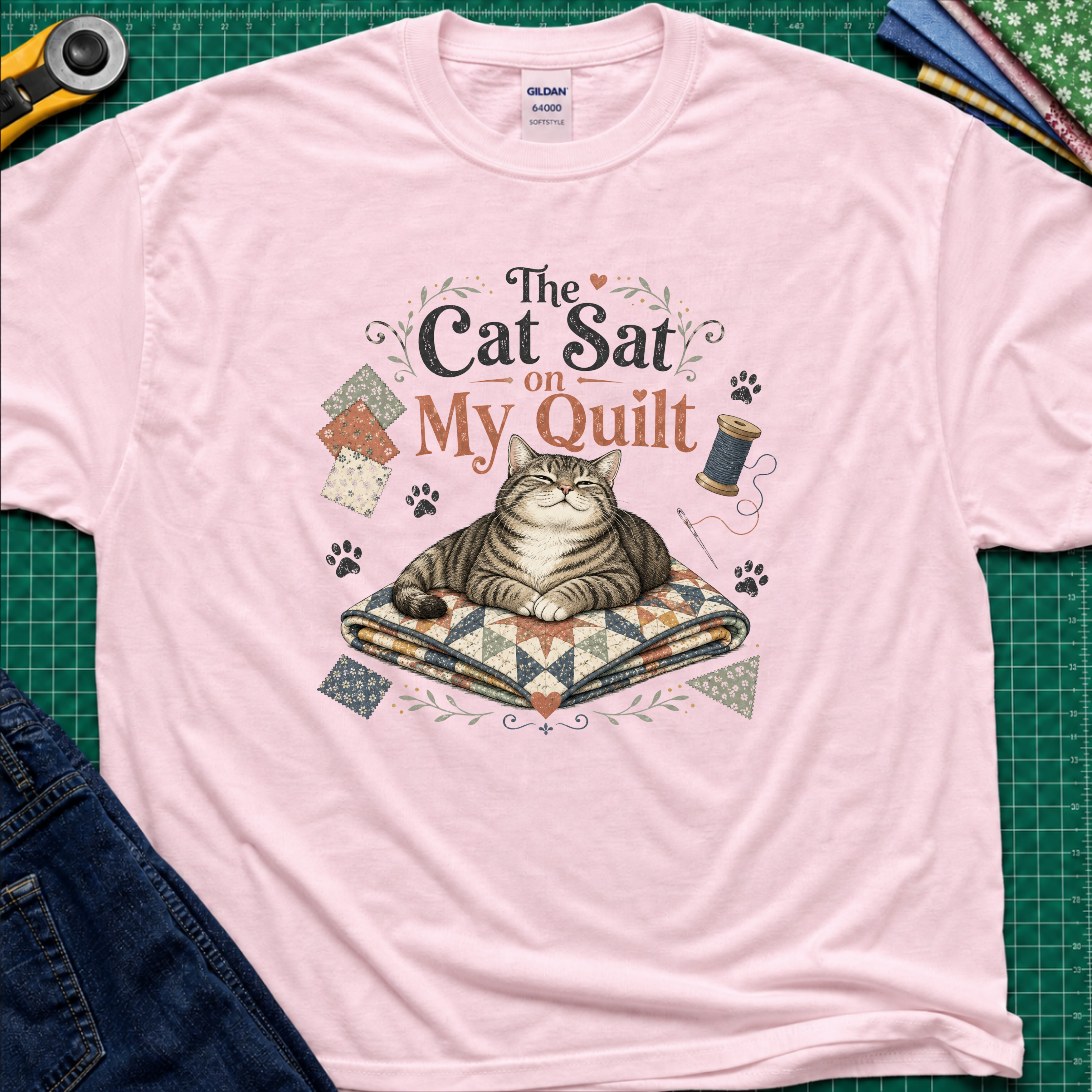 A light pink Gildan t-shirt laid flat on a green cutting mat, featuring a graphic of a tabby cat resting on a quilted blanket with the text 'The Cat Sat on My Quilt' with paw prints and fabric swatches.