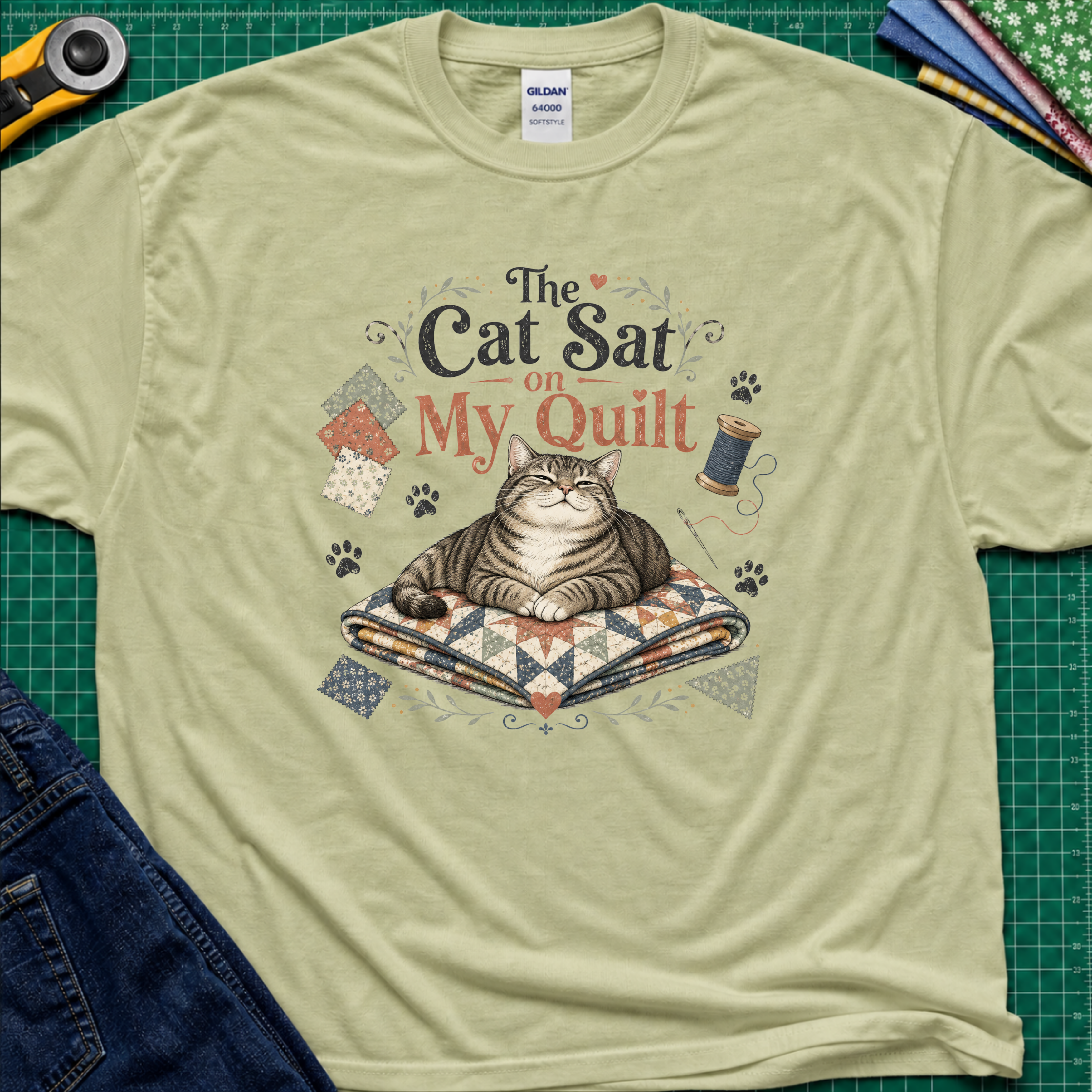 A light green Gildan t-shirt laid flat on a green cutting mat, featuring a graphic of a tabby cat resting on a quilted blanket with the text 'The Cat Sat on My Quilt' with paw prints and fabric swatches.