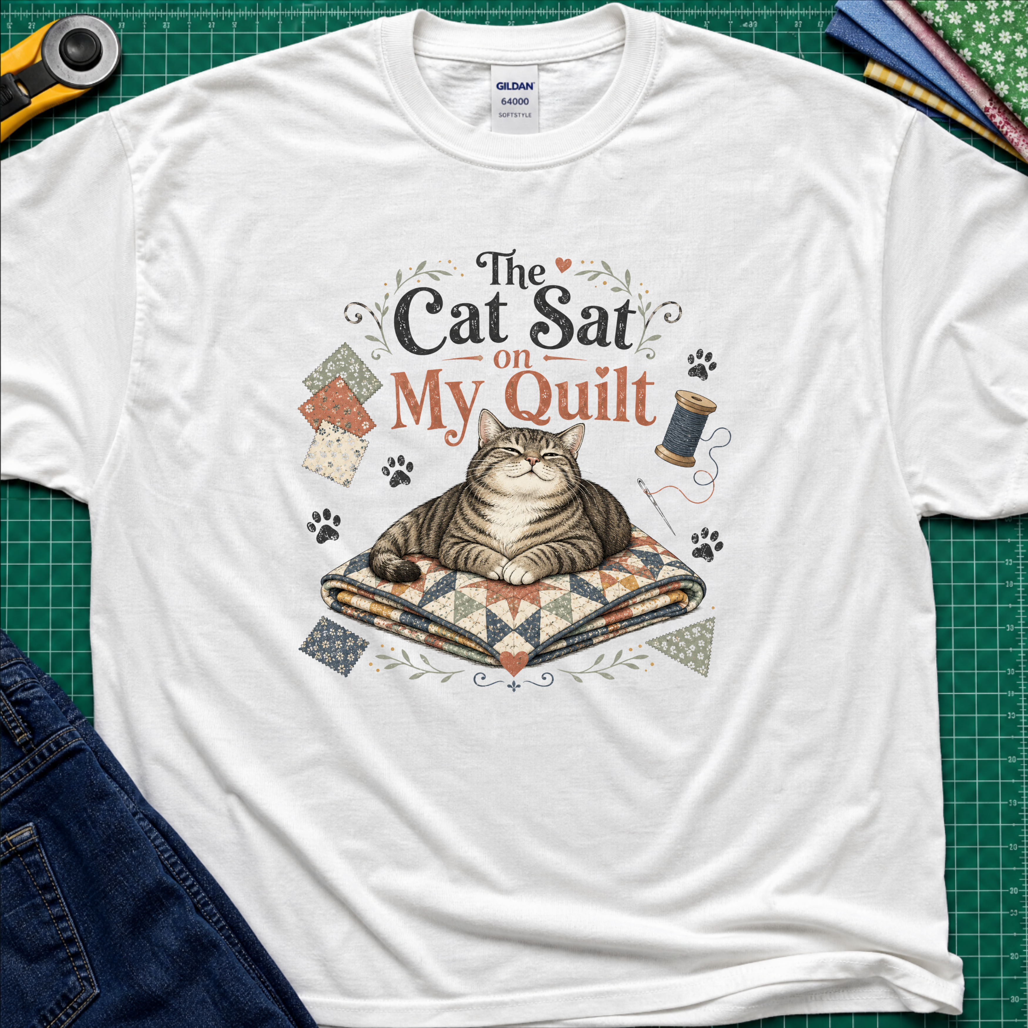 A white Gildan t-shirt laid flat on a green cutting mat, featuring a graphic of a tabby cat resting on a quilted blanket with the text 'The Cat Sat on My Quilt' with paw prints and fabric swatches.