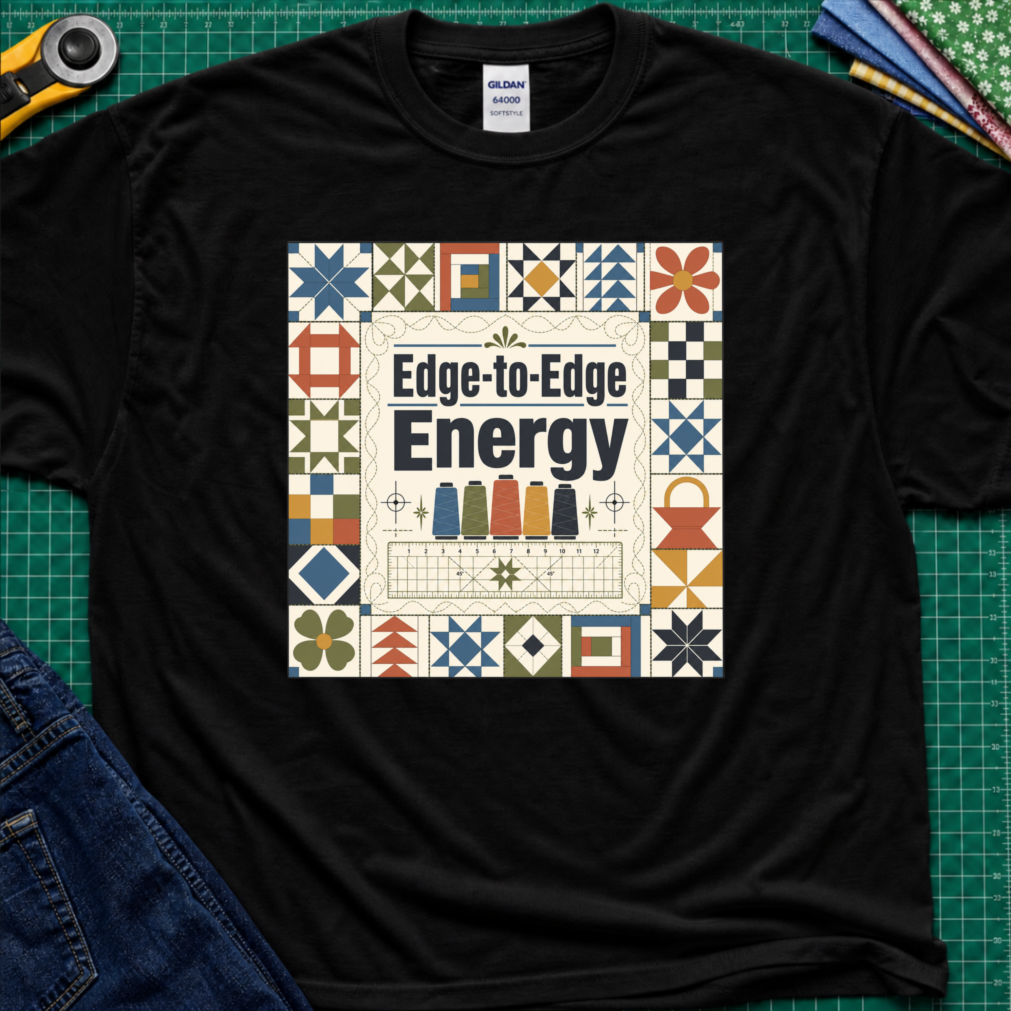 A black Gildan t-shirt featuring the text 'Edge-to-Edge Energy' in the center, surrounded by quilt blocks. The t-shirt lays flat on a green cutting mat with a rotary cutter, folded fabric stack and blue jeans partially visible in the corners.