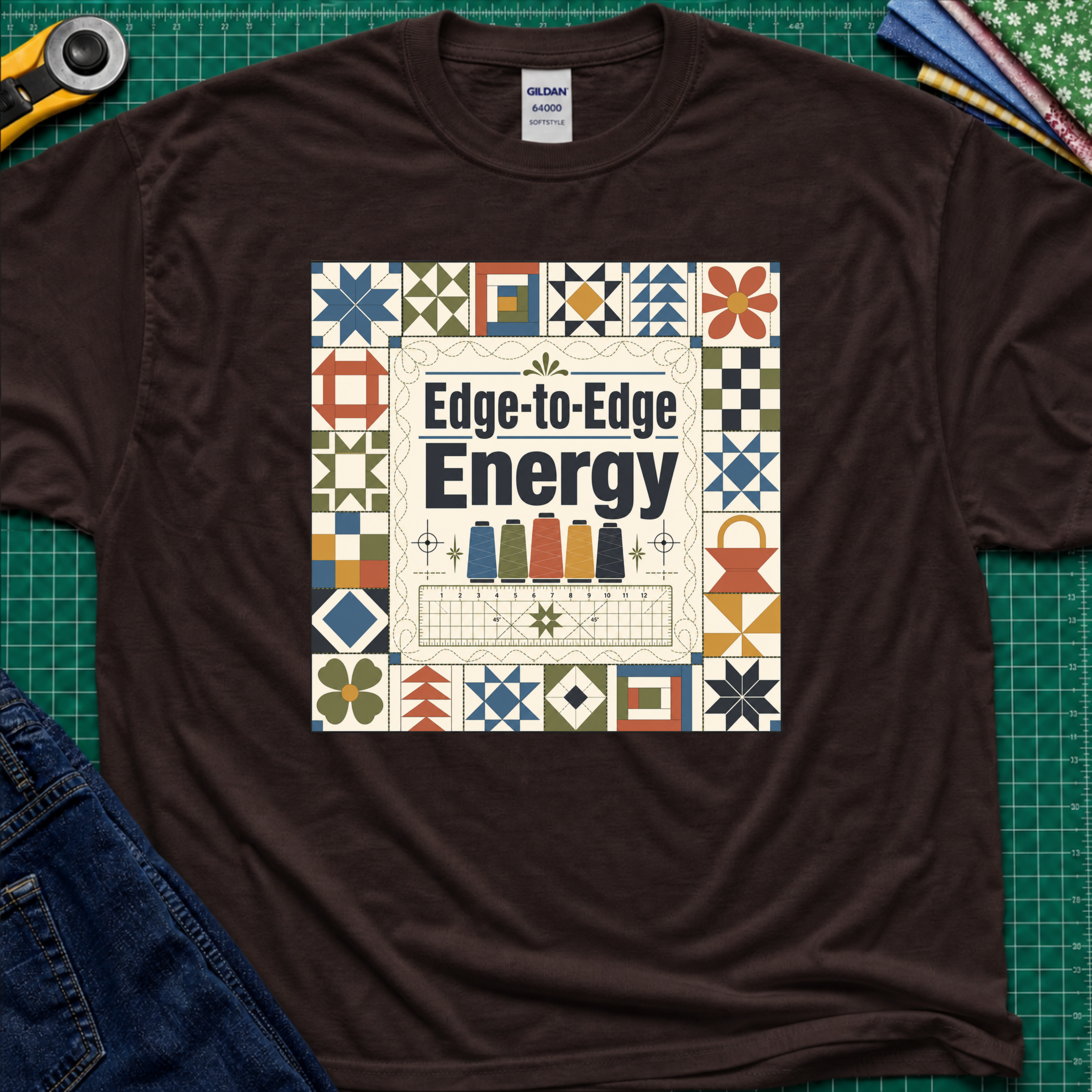 A dark chocolate Gildan t-shirt featuring the text 'Edge-to-Edge Energy' in the center, surrounded by quilt blocks. The t-shirt lays flat on a green cutting mat with a rotary cutter, folded fabric stack and blue jeans partially visible in the corners.