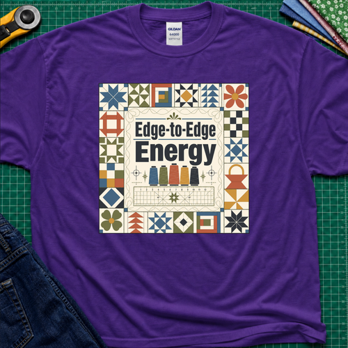 A purple Gildan t-shirt featuring the text 'Edge-to-Edge Energy' in the center, surrounded by quilt blocks. The t-shirt lays flat on a green cutting mat with a rotary cutter, folded fabric stack and blue jeans partially visible in the corners.