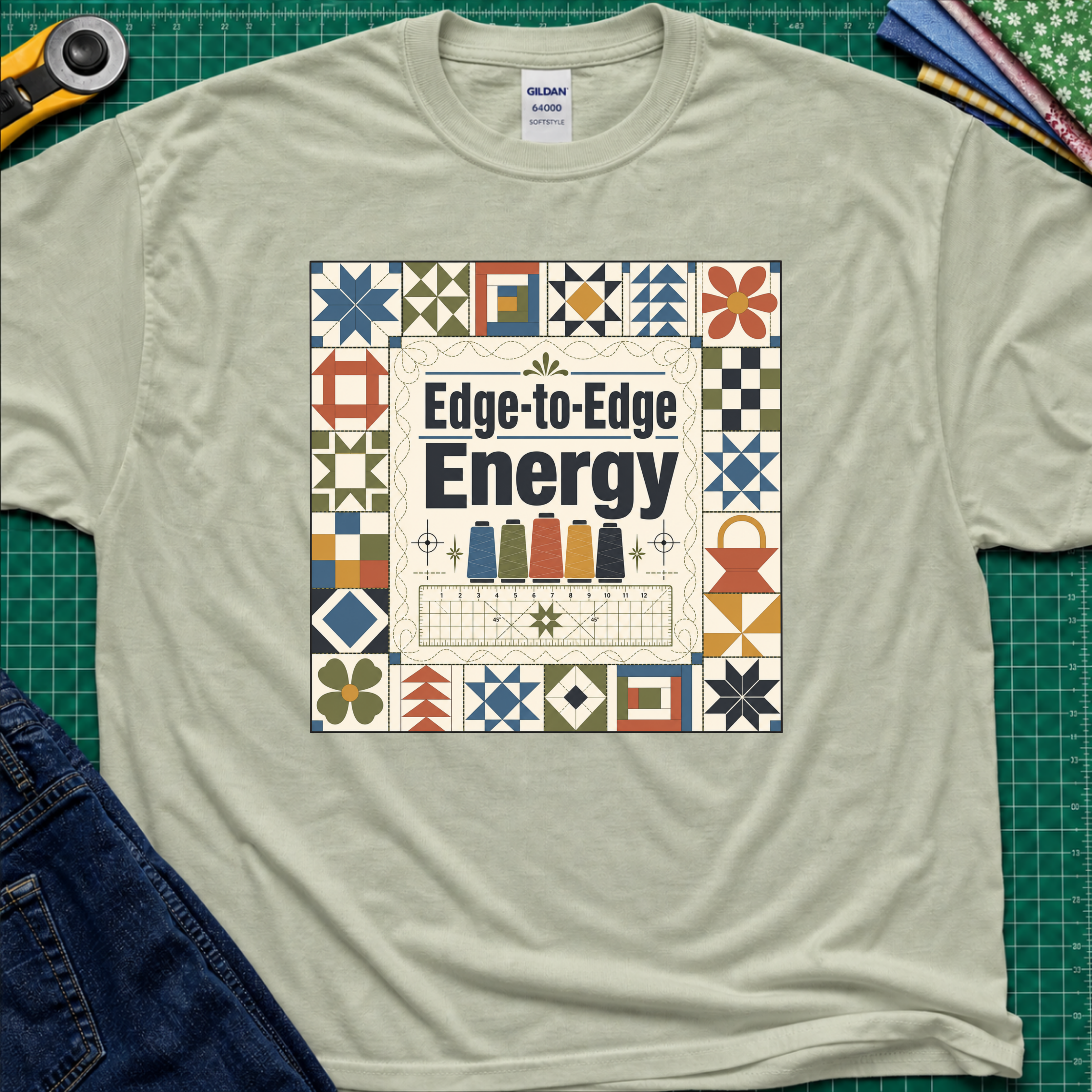 A light sage green Gildan t-shirt featuring the text 'Edge-to-Edge Energy' in the center, surrounded by quilt blocks. The t-shirt lays flat on a green cutting mat with a rotary cutter, folded fabric stack and blue jeans partially visible in the corners.