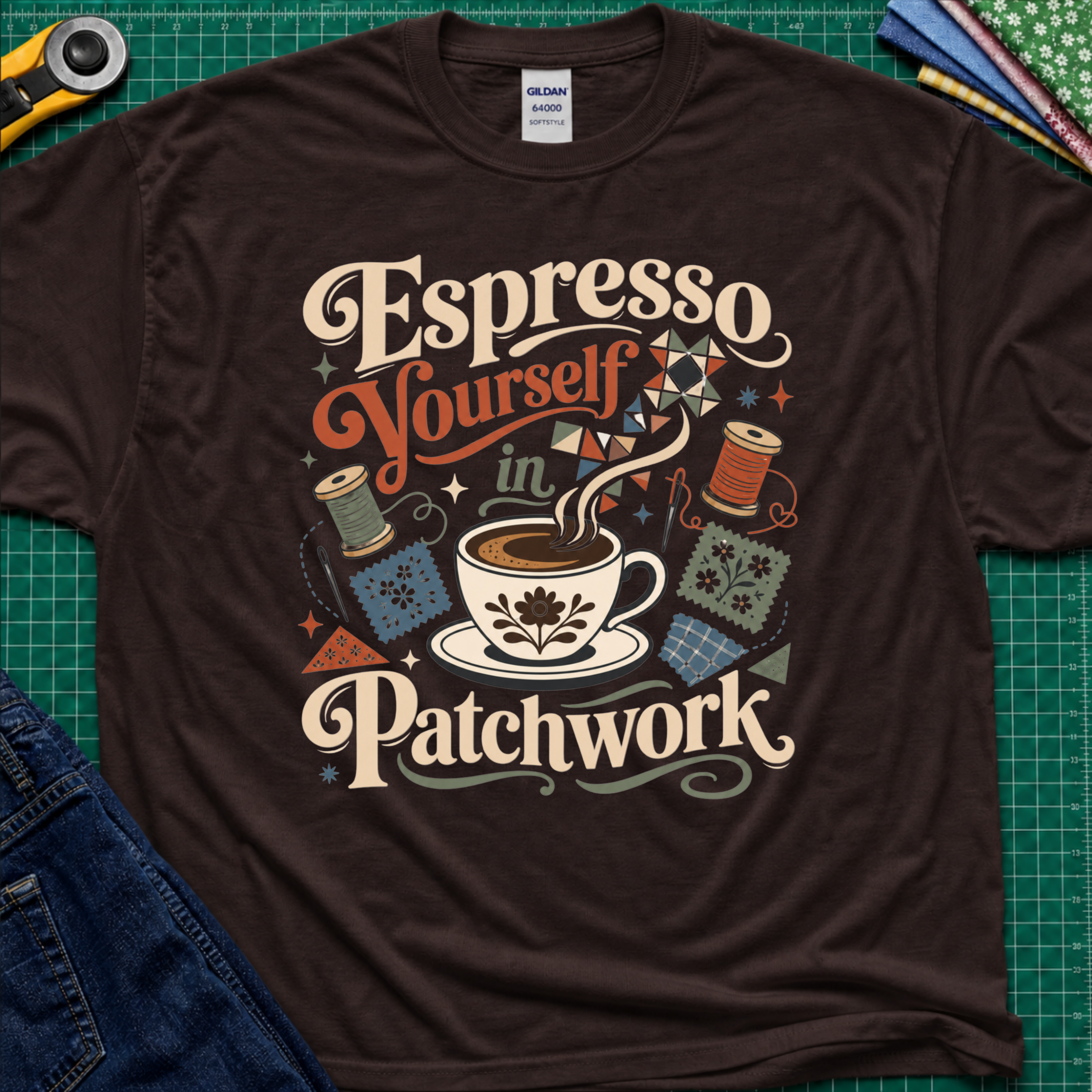 An dark chocolate Gildan t-shirt with a colorful retro graphic that reads 'Espresso Yourself in Patchwork' around an illustration of a coffee cup, fabric swatches and cotton reels. It is laid flat on a green cutting mat with a rotary cutter, fabric stack and blue jeans partially visible in the corners.