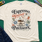 An light cream Gildan t-shirt with a colorful retro graphic that reads 'Espresso Yourself in Patchwork' around an illustration of a coffee cup, fabric swatches and cotton reels. It is laid flat on a green cutting mat with a rotary cutter, fabric stack and blue jeans partially visible in the corners.