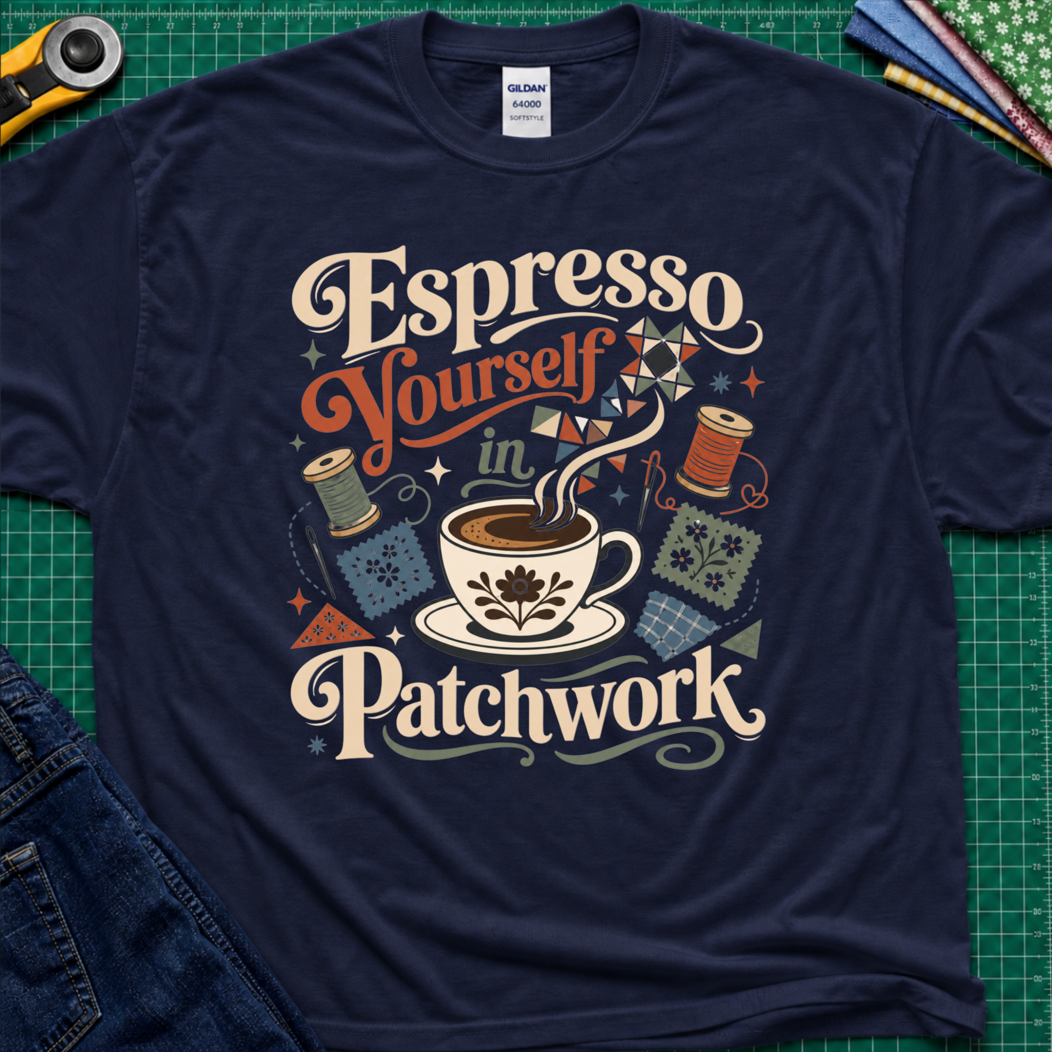 An navy blue Gildan t-shirt with a colorful retro graphic that reads 'Espresso Yourself in Patchwork' around an illustration of a coffee cup, fabric swatches and cotton reels. It is laid flat on a green cutting mat with a rotary cutter, fabric stack and blue jeans partially visible in the corners.
