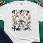 An white Gildan t-shirt with a colorful retro graphic that reads 'Espresso Yourself in Patchwork' around an illustration of a coffee cup, fabric swatches and cotton reels. It is laid flat on a green cutting mat with a rotary cutter, fabric stack and blue jeans partially visible in the corners.