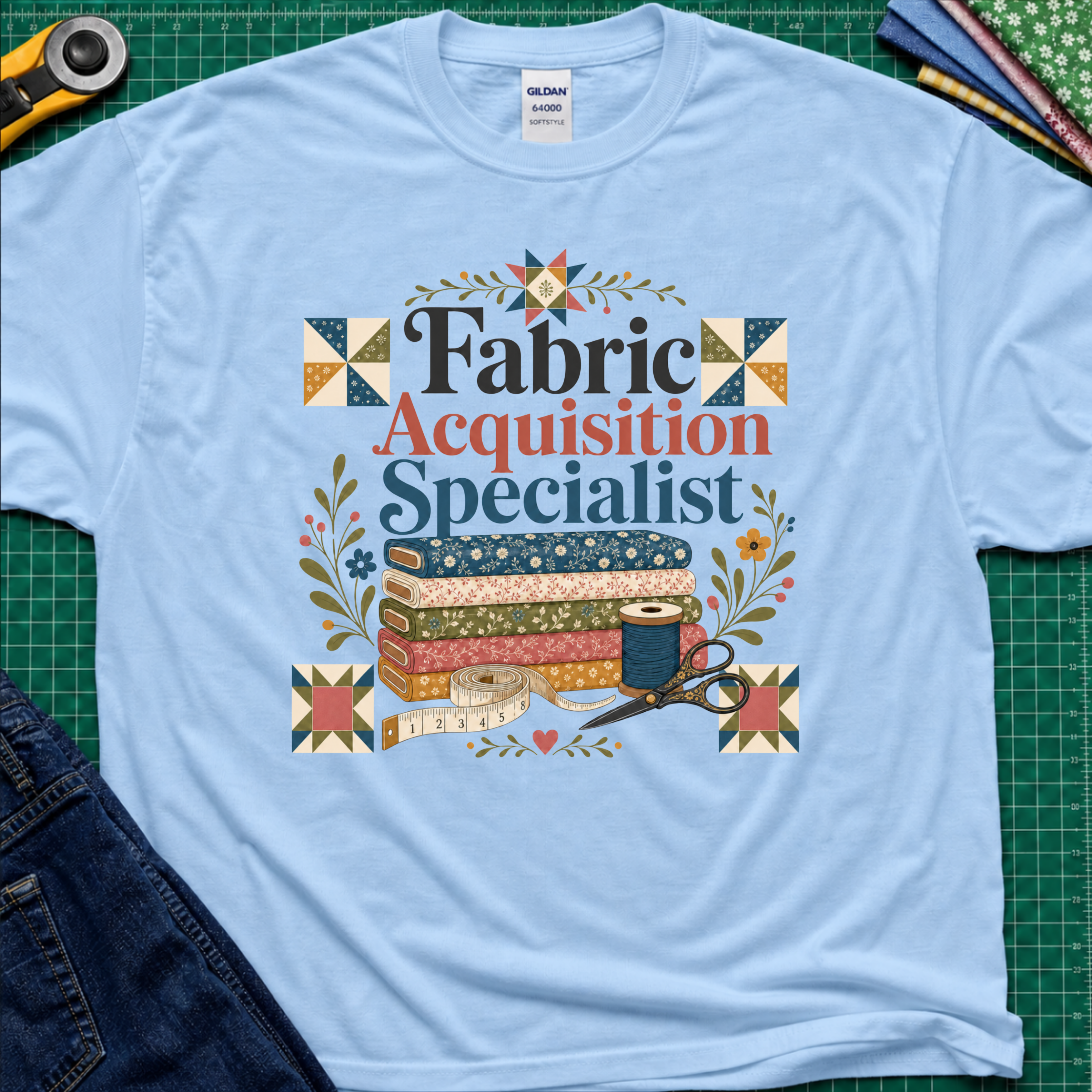 A light blue Gildan t-shirt with a colorful graphic that reads 'Fabric Acquisition Specialist' surrounded by quilting motifs like fabrics, a spool, scissors, and flowers. The shirt is laid flat on a green cutting mat with sewing tools and jeans partially visible around it.
