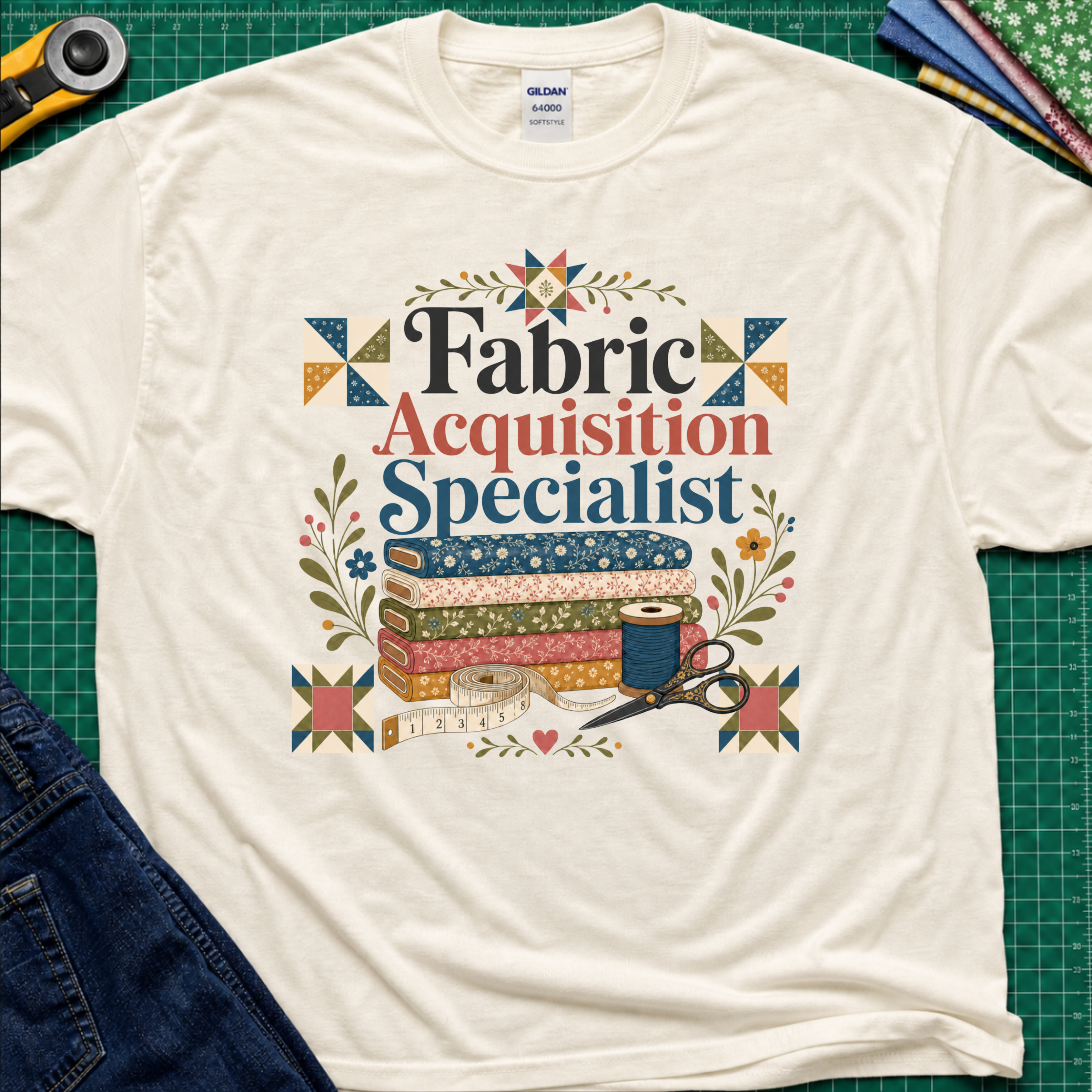 A light cream Gildan t-shirt with a colorful graphic that reads 'Fabric Acquisition Specialist' surrounded by quilting motifs like fabrics, a spool, scissors, and flowers. The shirt is laid flat on a green cutting mat with sewing tools and jeans partially visible around it.