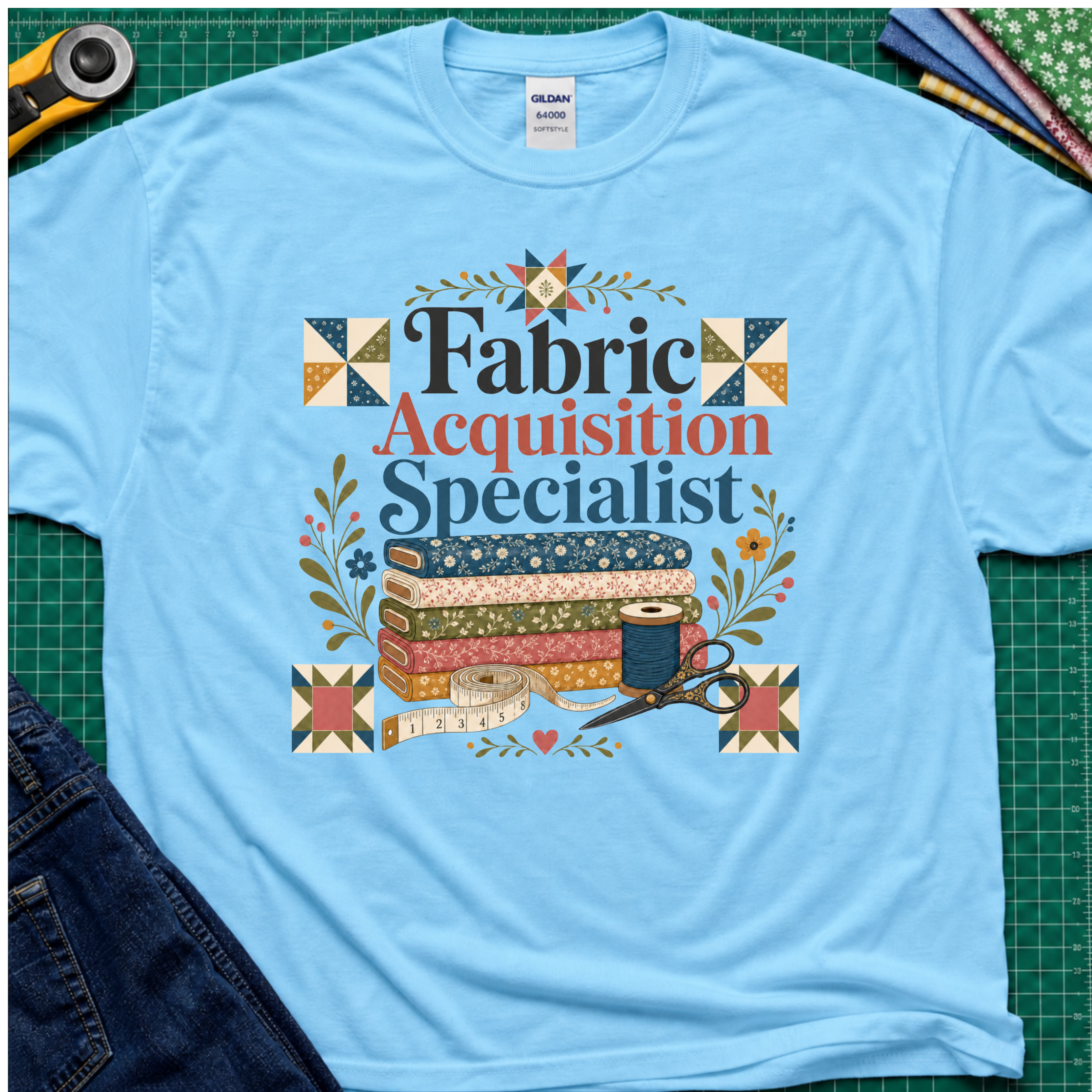 A sky blue Gildan t-shirt with a colorful graphic that reads 'Fabric Acquisition Specialist' surrounded by quilting motifs like fabrics, a spool, scissors, and flowers. The shirt is laid flat on a green cutting mat with sewing tools and jeans partially visible around it.