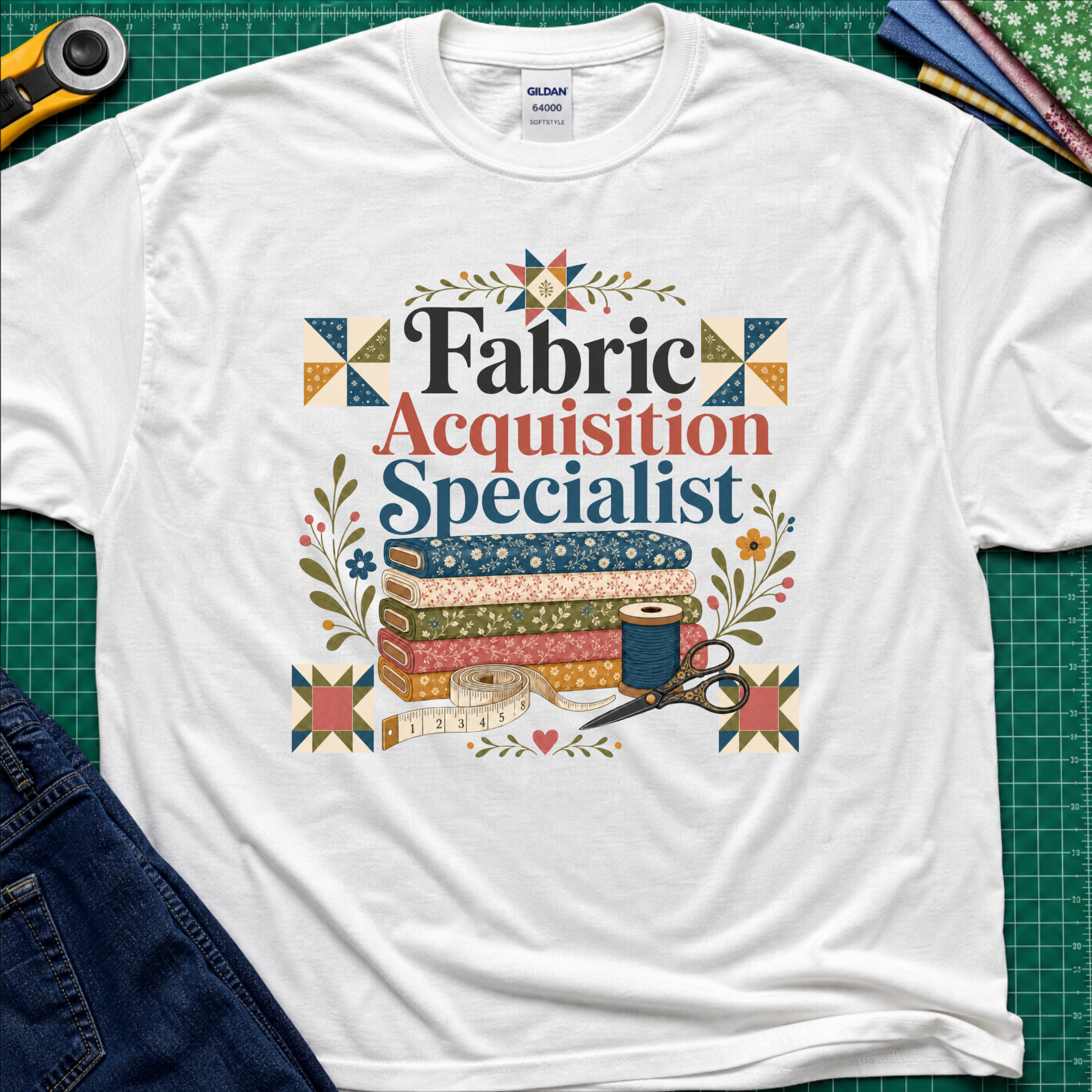 A white Gildan t-shirt with a colorful graphic that reads 'Fabric Acquisition Specialist' surrounded by quilting motifs like fabrics, a spool, scissors, and flowers. The shirt is laid flat on a green cutting mat with sewing tools and jeans partially visible around it.