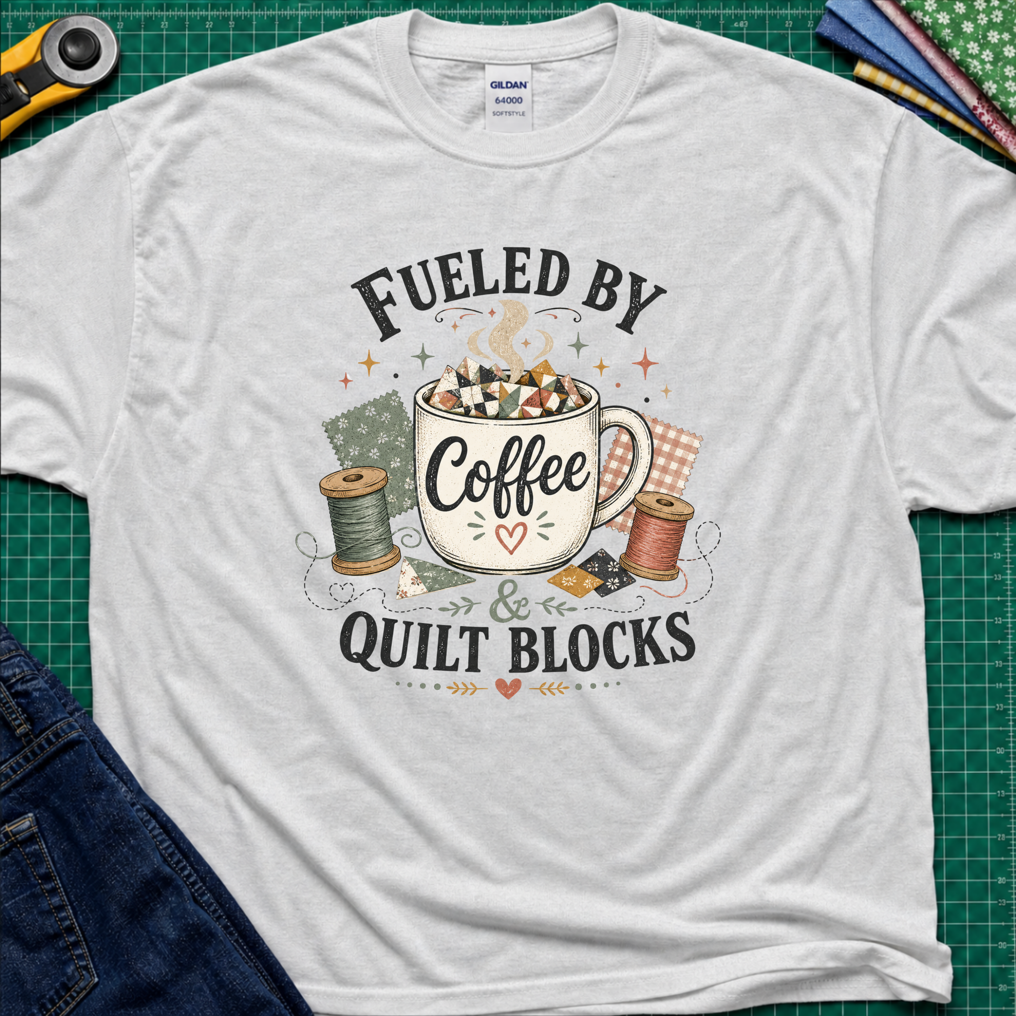 A light grey Gildan t-shirt with a graphic that reads 'FUELED BY Coffee & QUILT BLOCKS' surrounding an illustration of a coffee mug filled with quilt blocks, styled on a green cutting mat with sewing supplies.