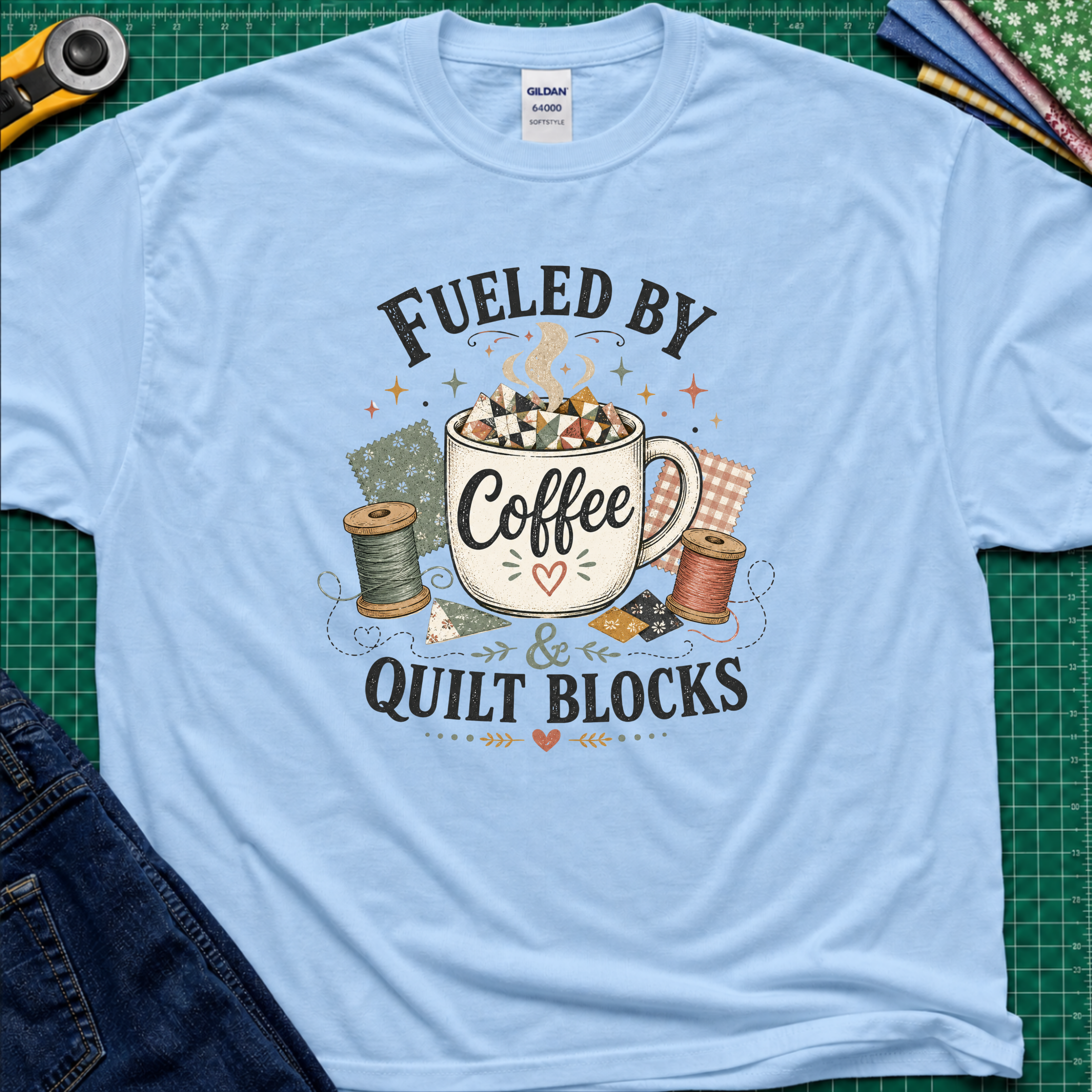 A light blue Gildan t-shirt with a graphic that reads 'FUELED BY Coffee & QUILT BLOCKS' surrounding an illustration of a coffee mug filled with quilt blocks, styled on a green cutting mat with sewing supplies.