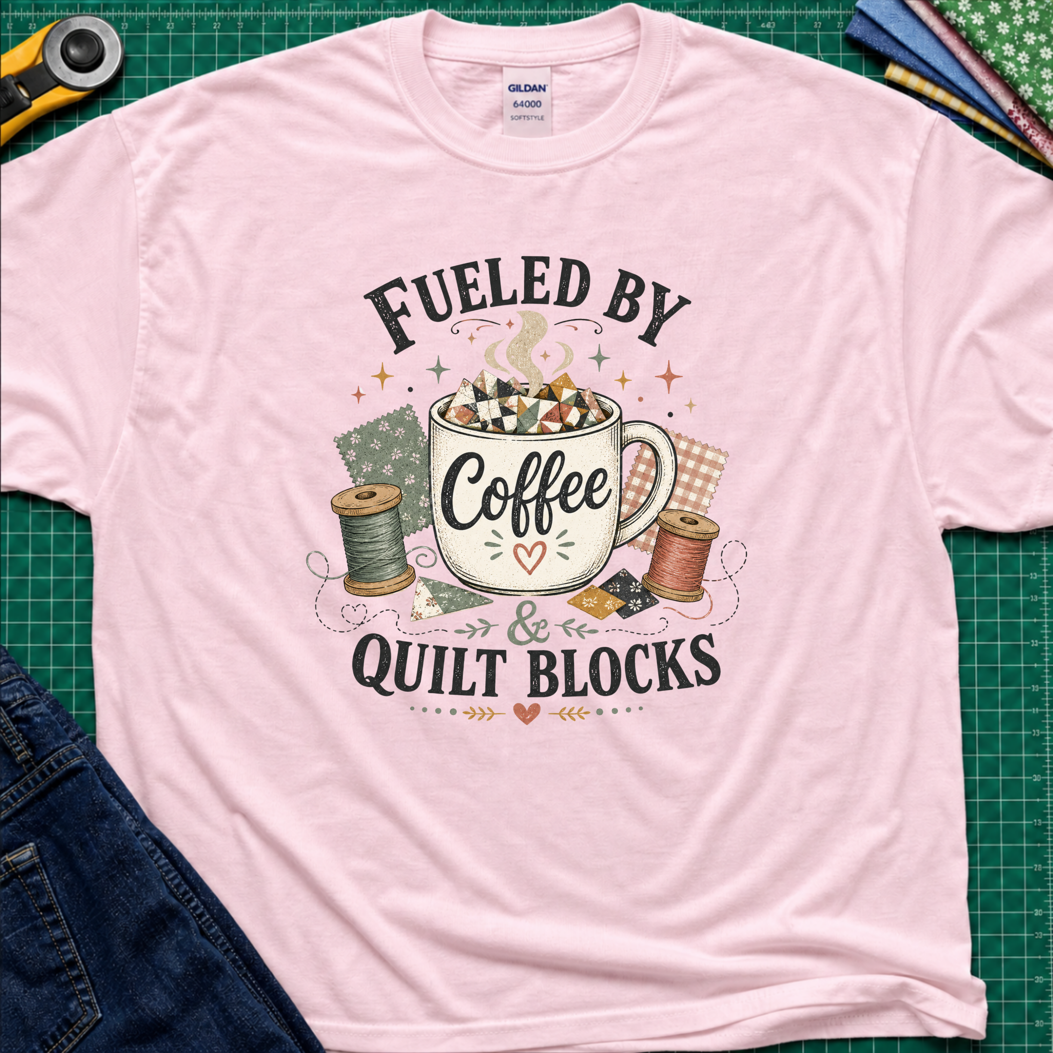 A light pink Gildan t-shirt with a graphic that reads 'FUELED BY Coffee & QUILT BLOCKS' surrounding an illustration of a coffee mug filled with quilt blocks, styled on a green cutting mat with sewing supplies.