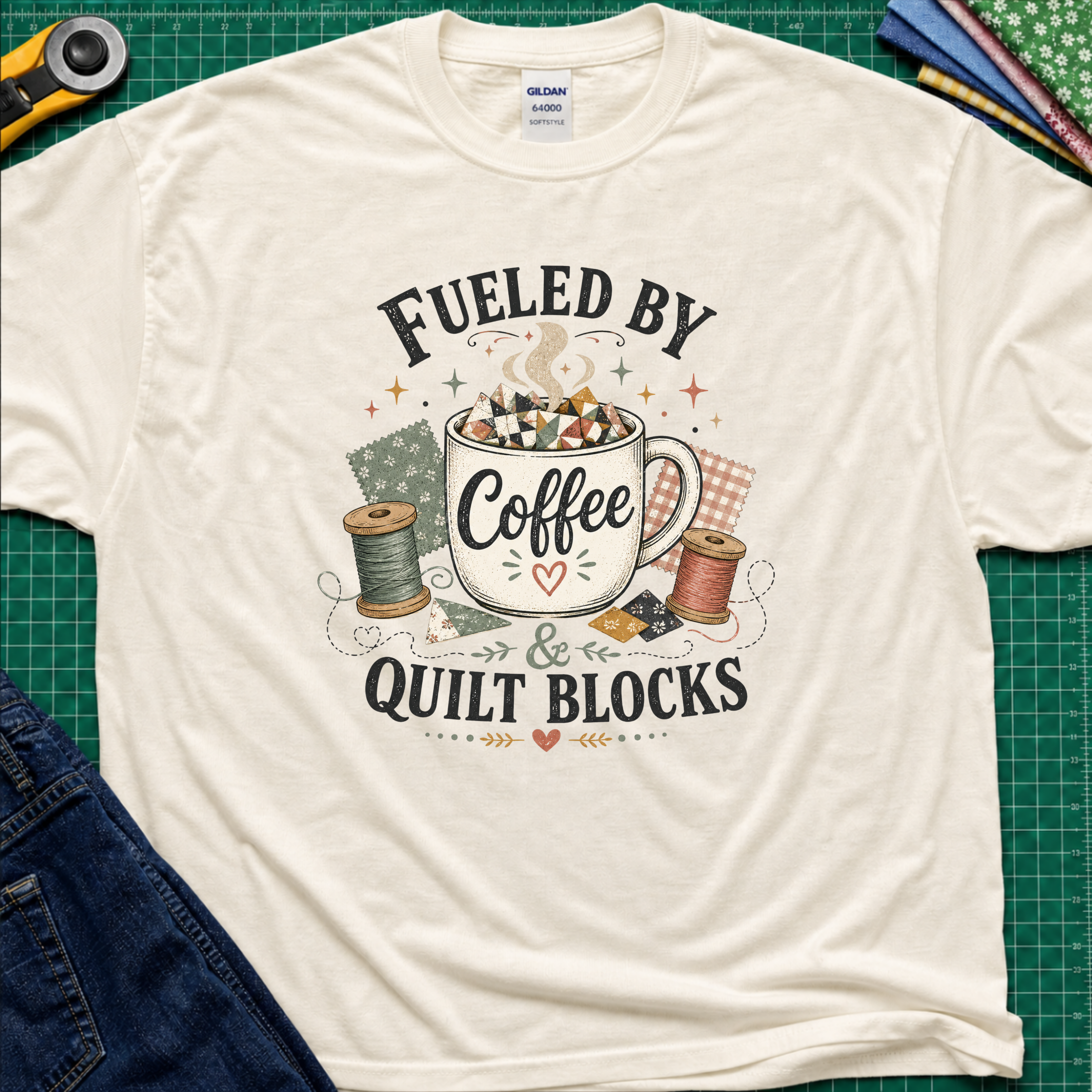 A light cream Gildan t-shirt with a graphic that reads 'FUELED BY Coffee & QUILT BLOCKS' surrounding an illustration of a coffee mug filled with quilt blocks, styled on a green cutting mat with sewing supplies.