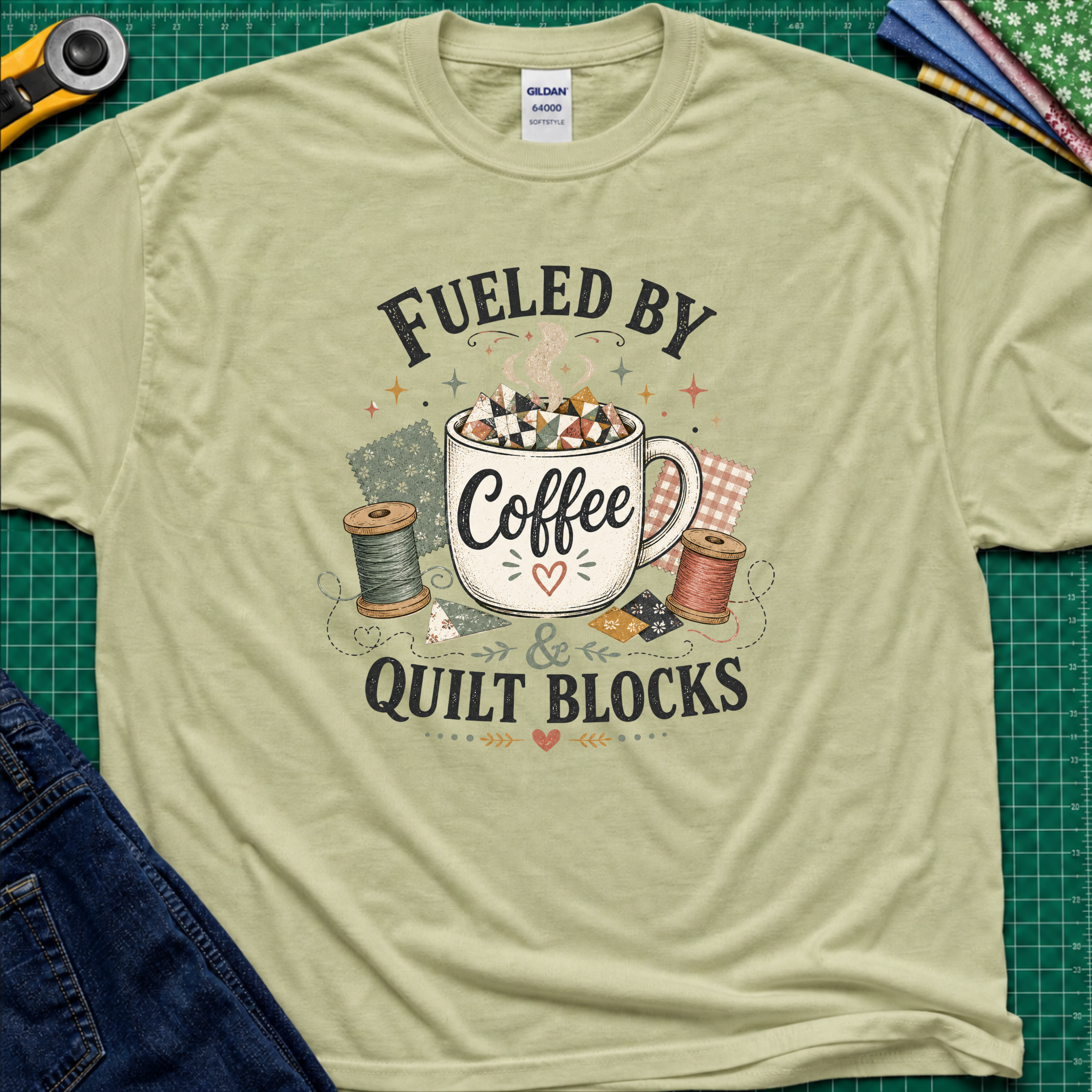 A light pistachio green Gildan t-shirt with a graphic that reads 'FUELED BY Coffee & QUILT BLOCKS' surrounding an illustration of a coffee mug filled with quilt blocks, styled on a green cutting mat with sewing supplies.