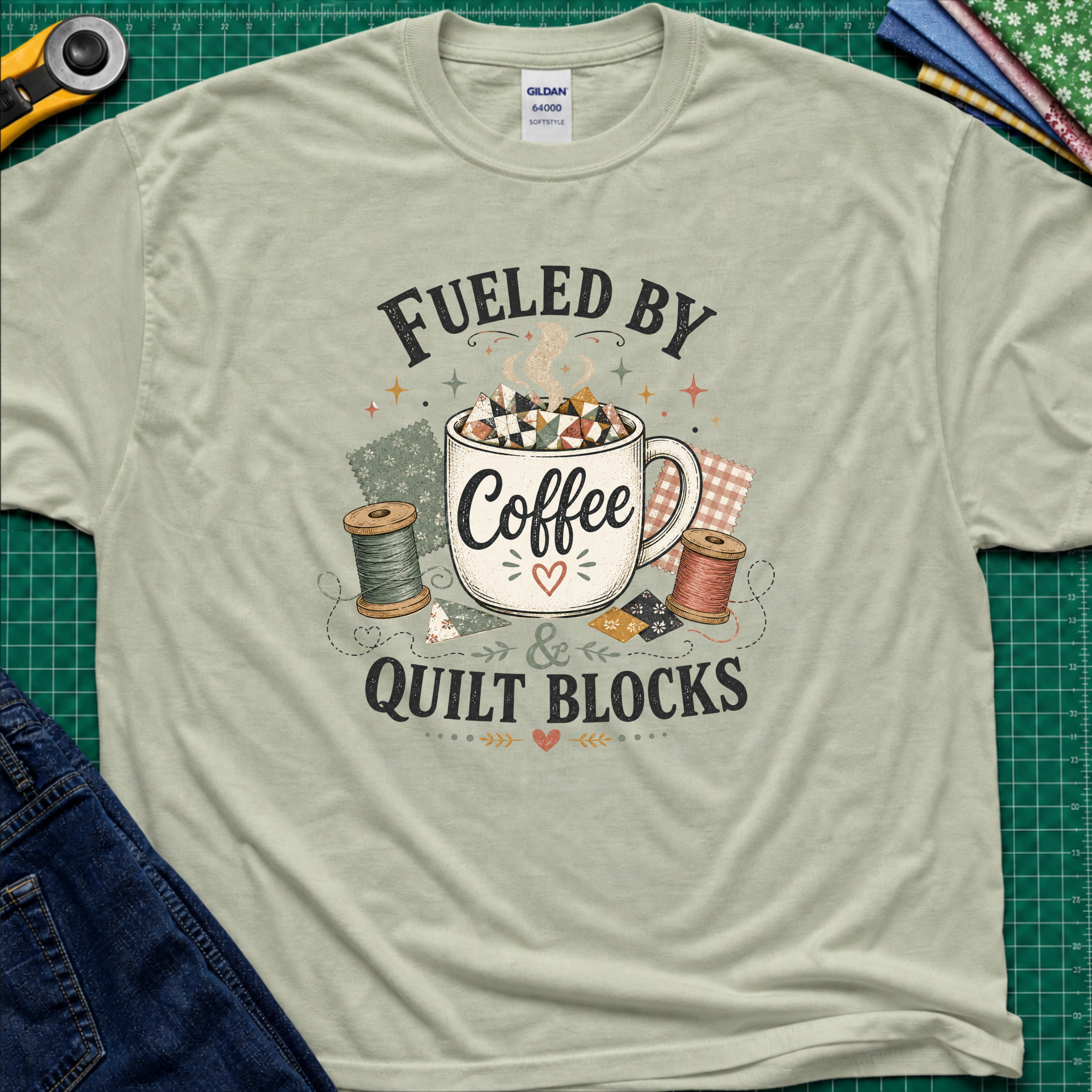 A light sage green Gildan t-shirt with a graphic that reads 'FUELED BY Coffee & QUILT BLOCKS' surrounding an illustration of a coffee mug filled with quilt blocks, styled on a green cutting mat with sewing supplies.
