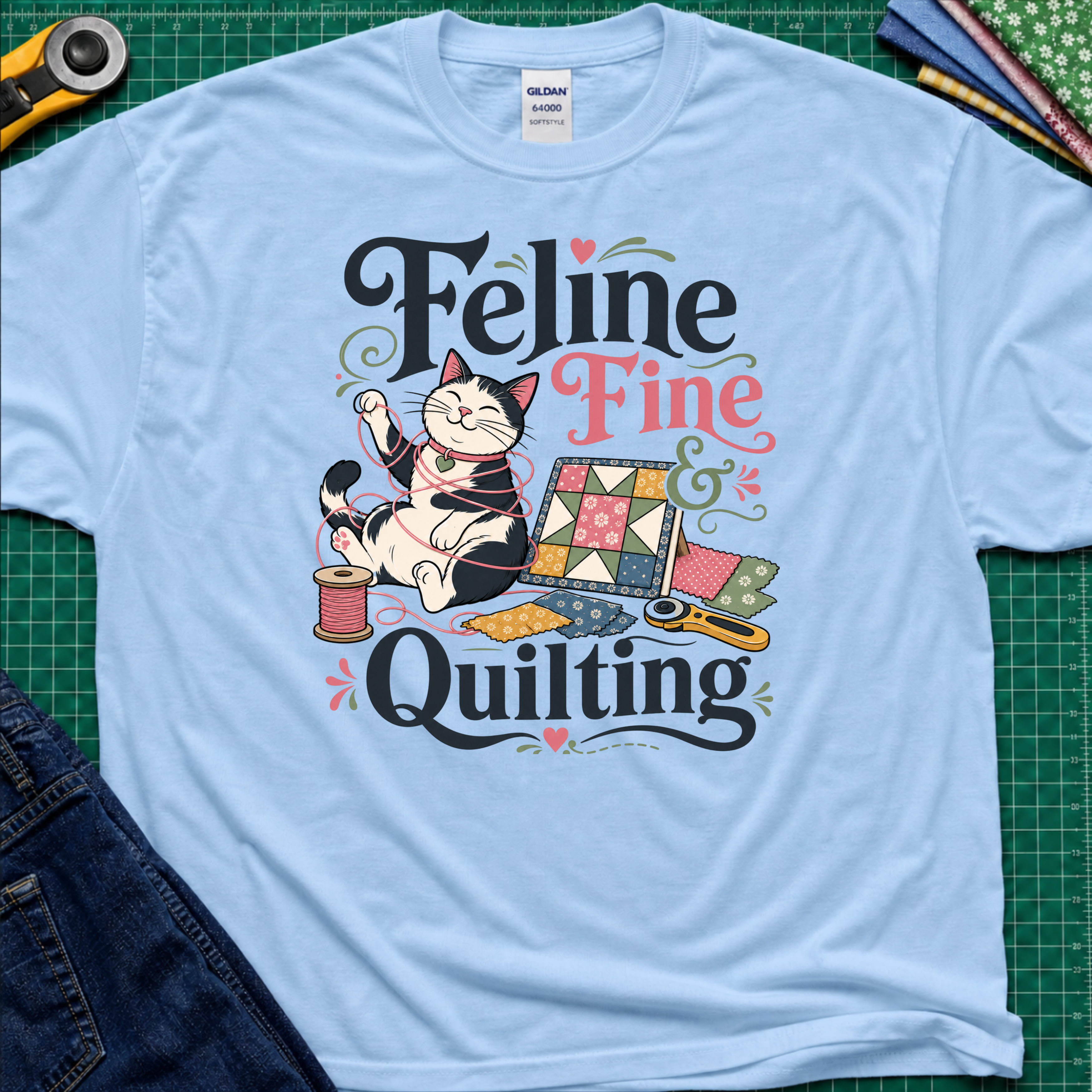 A light blue Gildan t-shirt with a colorful graphic that reads 'Feline Fine Quilting' featuring a cartoon cat and quilting supplies, laid flat on a green cutting mat with crafting props and blue jeans partially visible.