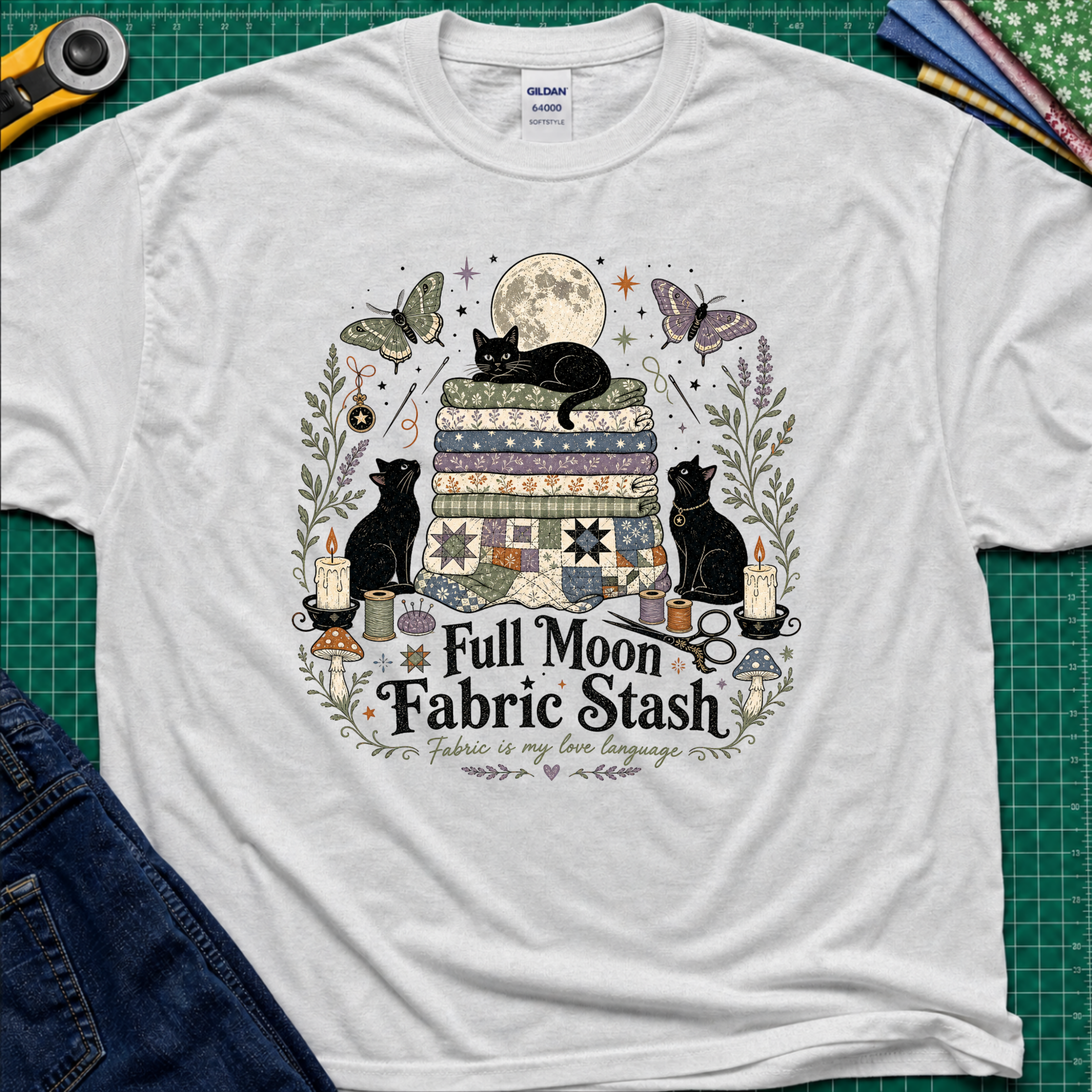 A light grey Gildan t-shirt with a detailed graphic print of three black cats, butterflies, quilts, and sewing tools under a full moon, with the text 'Full Moon Fabric Stash'. The shirt is laid flat on a green cutting mat with crafting tools and jeans partially visible around it.