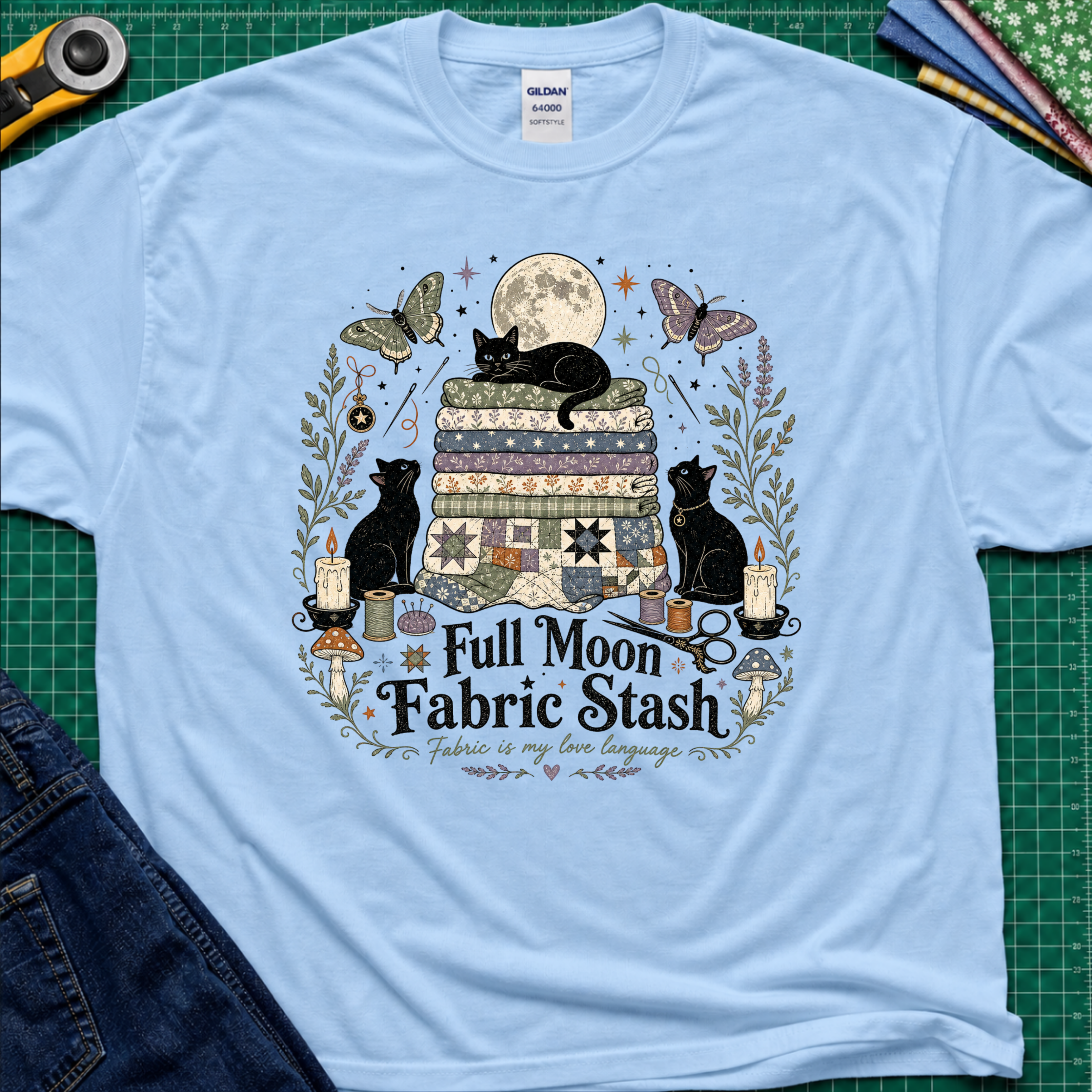 A light blue Gildan t-shirt with a detailed graphic print of three black cats, butterflies, quilts, and sewing tools under a full moon, with the text 'Full Moon Fabric Stash'. The shirt is laid flat on a green cutting mat with crafting tools and jeans partially visible around it.
