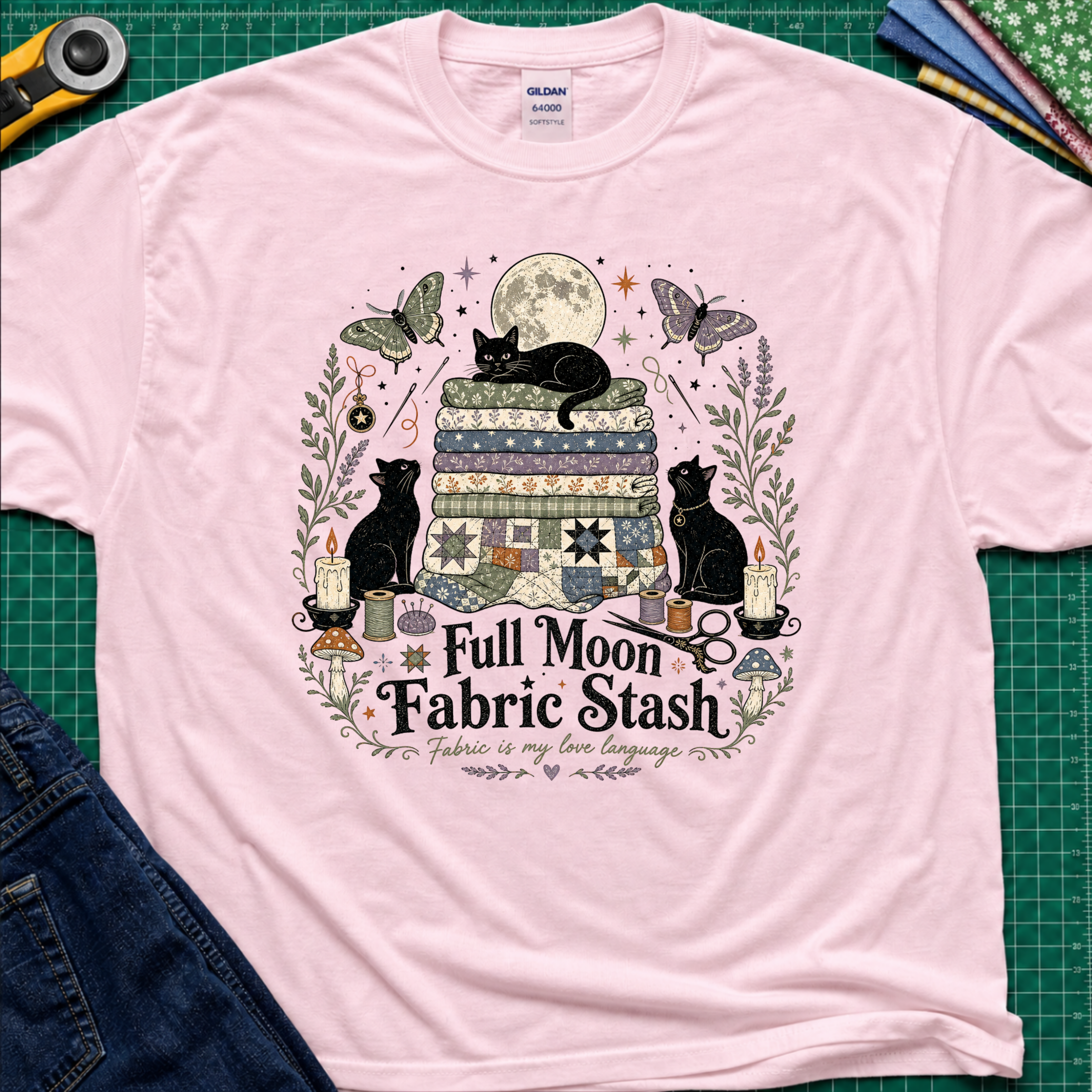 A light pink Gildan t-shirt with a detailed graphic print of three black cats, butterflies, quilts, and sewing tools under a full moon, with the text 'Full Moon Fabric Stash'. The shirt is laid flat on a green cutting mat with crafting tools and jeans partially visible around it.
