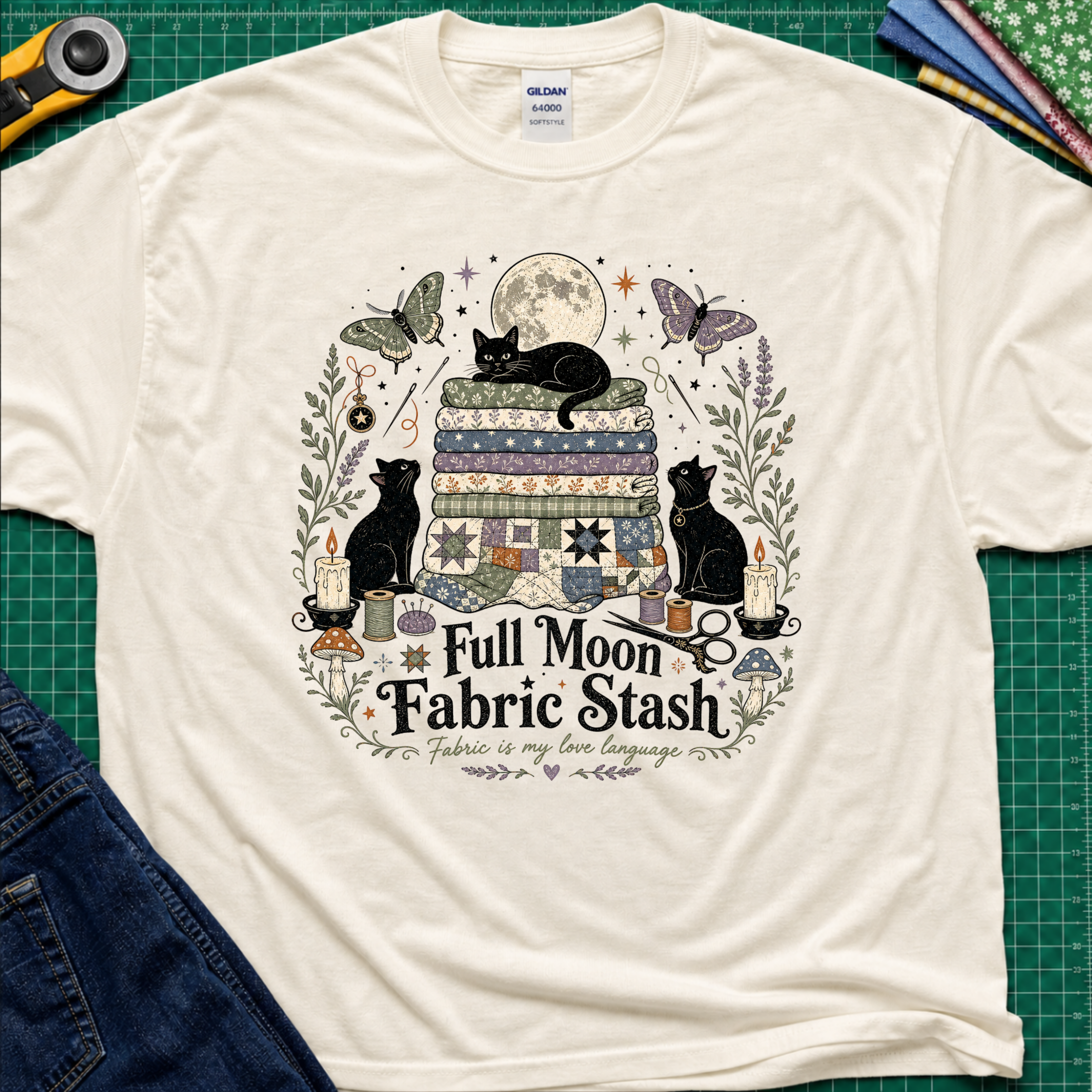 A light cream Gildan t-shirt with a detailed graphic print of three black cats, butterflies, quilts, and sewing tools under a full moon, with the text 'Full Moon Fabric Stash'. The shirt is laid flat on a green cutting mat with crafting tools and jeans partially visible around it.