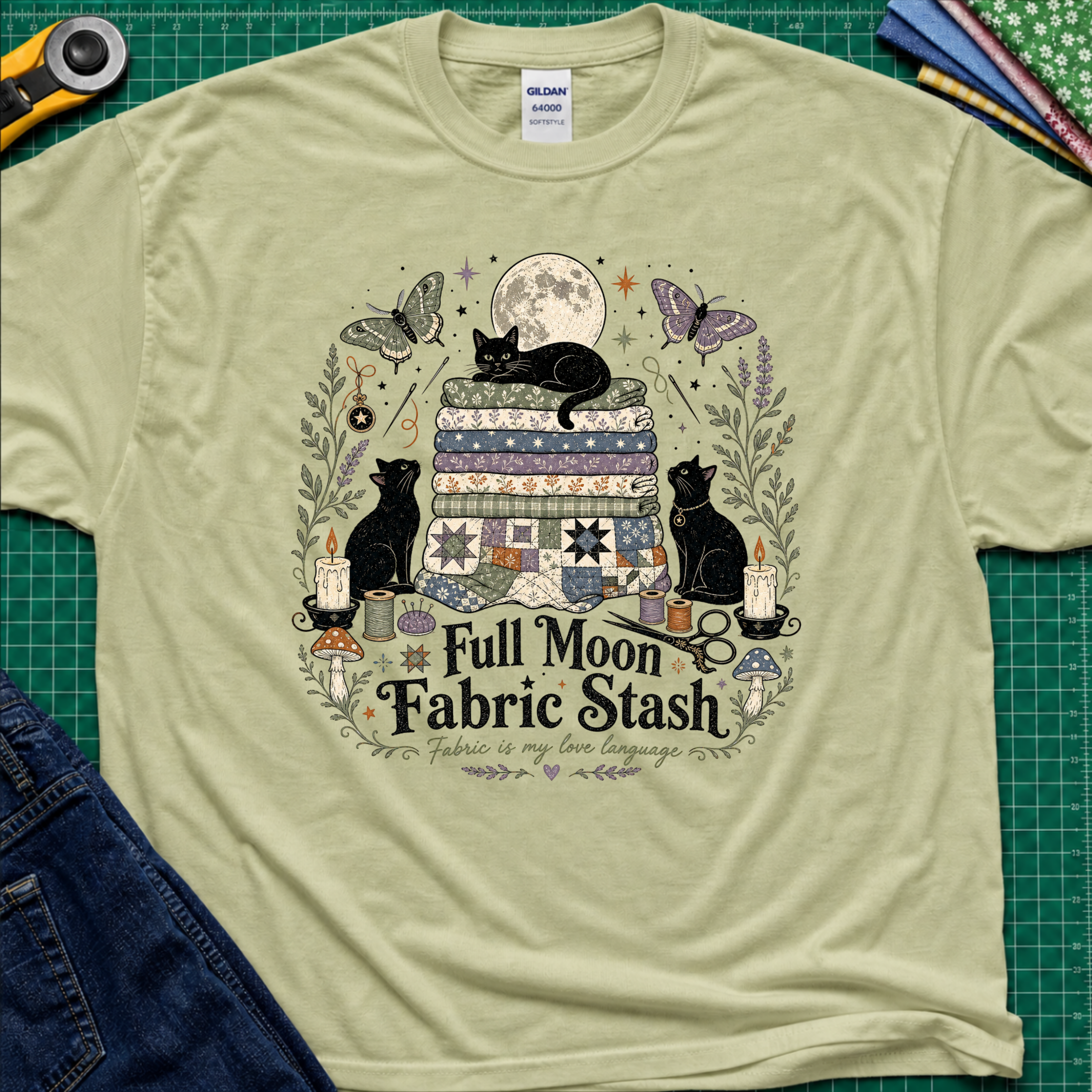 A light pistachio green Gildan t-shirt with a detailed graphic print of three black cats, butterflies, quilts, and sewing tools under a full moon, with the text 'Full Moon Fabric Stash'. The shirt is laid flat on a green cutting mat with crafting tools and jeans partially visible around it.