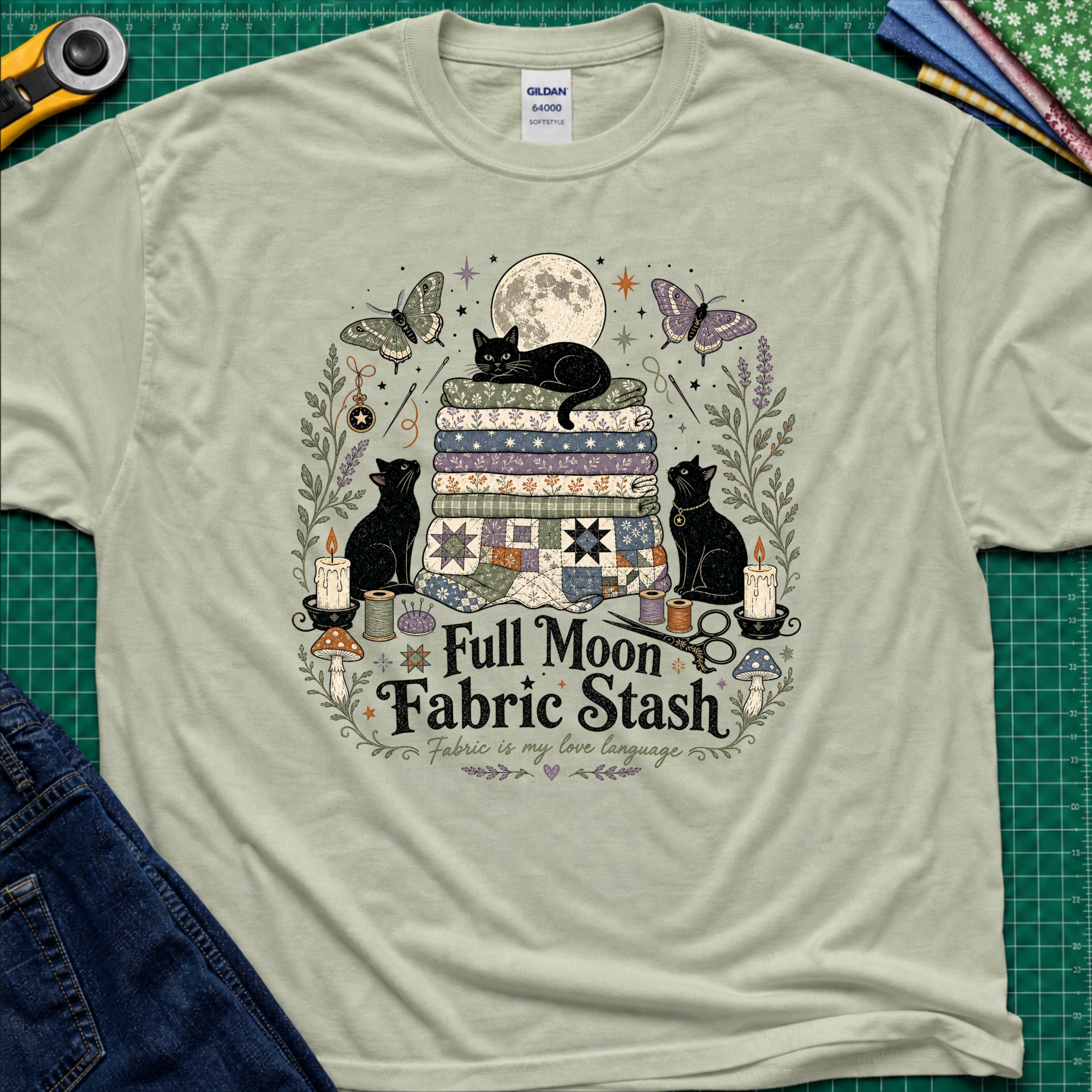 A light sage green Gildan t-shirt with a detailed graphic print of three black cats, butterflies, quilts, and sewing tools under a full moon, with the text 'Full Moon Fabric Stash'. The shirt is laid flat on a green cutting mat with crafting tools and jeans partially visible around it.