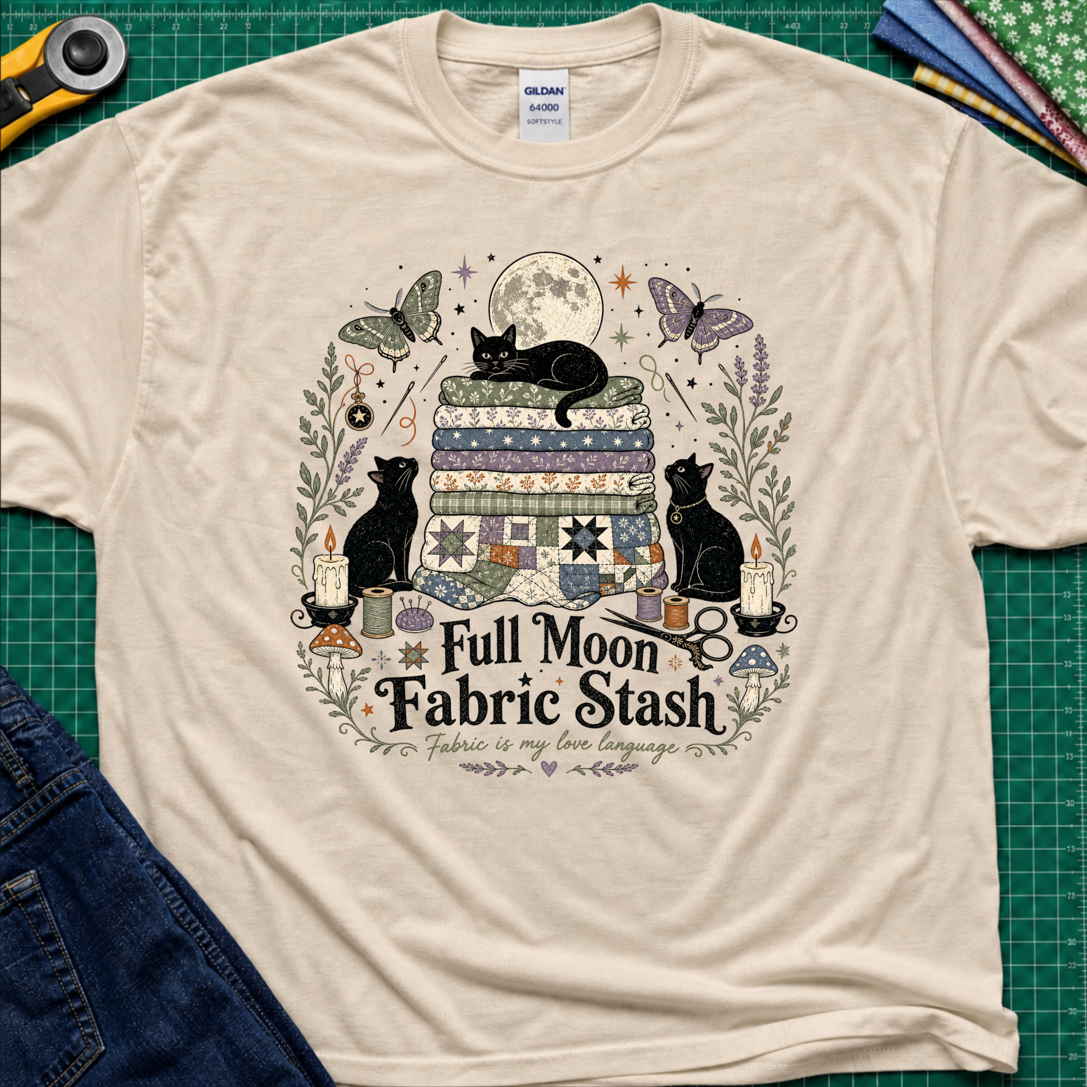 A dark cream Gildan t-shirt with a detailed graphic print of three black cats, butterflies, quilts, and sewing tools under a full moon, with the text 'Full Moon Fabric Stash'. The shirt is laid flat on a green cutting mat with crafting tools and jeans partially visible around it.