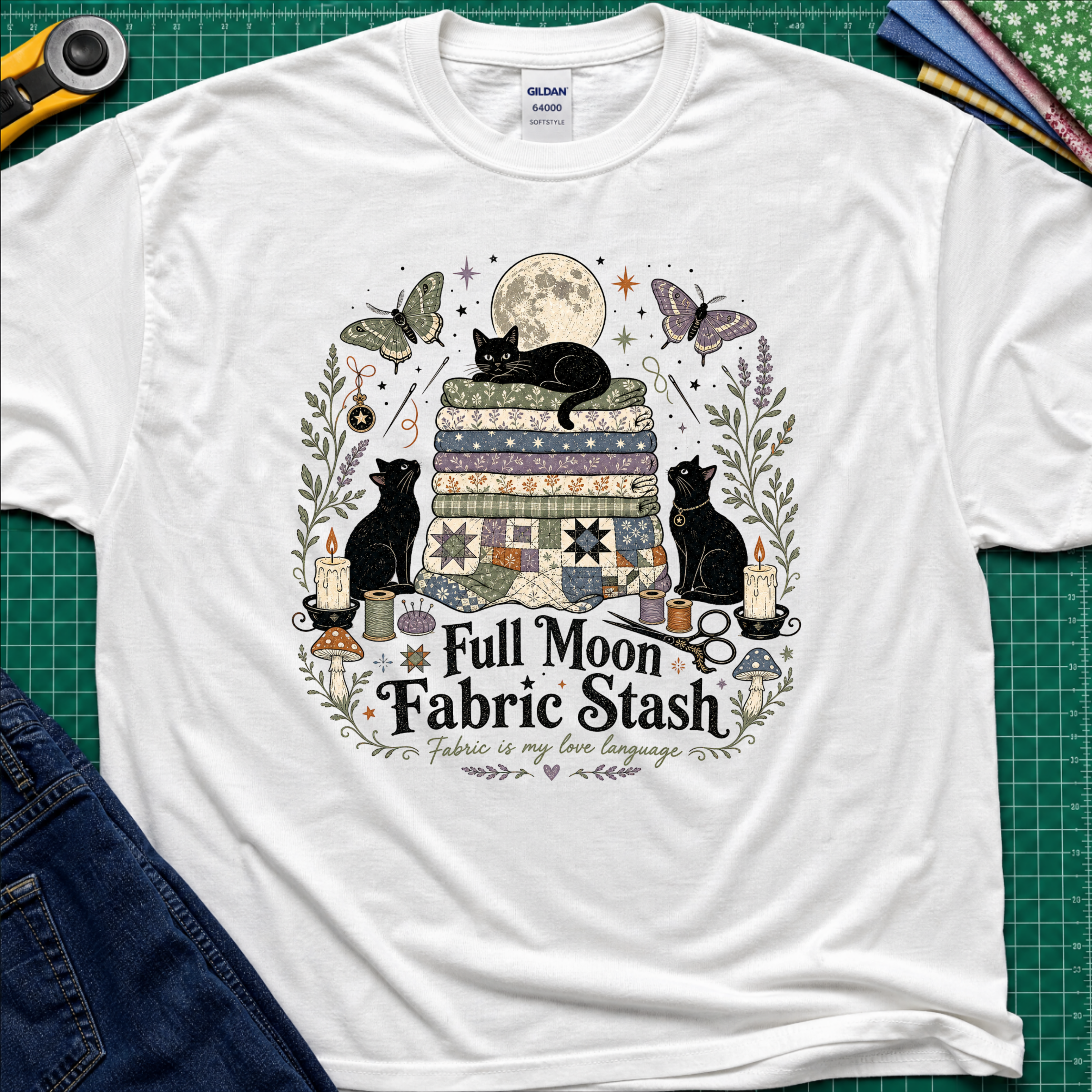 A white Gildan t-shirt with a detailed graphic print of three black cats, butterflies, quilts, and sewing tools under a full moon, with the text 'Full Moon Fabric Stash'. The shirt is laid flat on a green cutting mat with crafting tools and jeans partially visible around it.