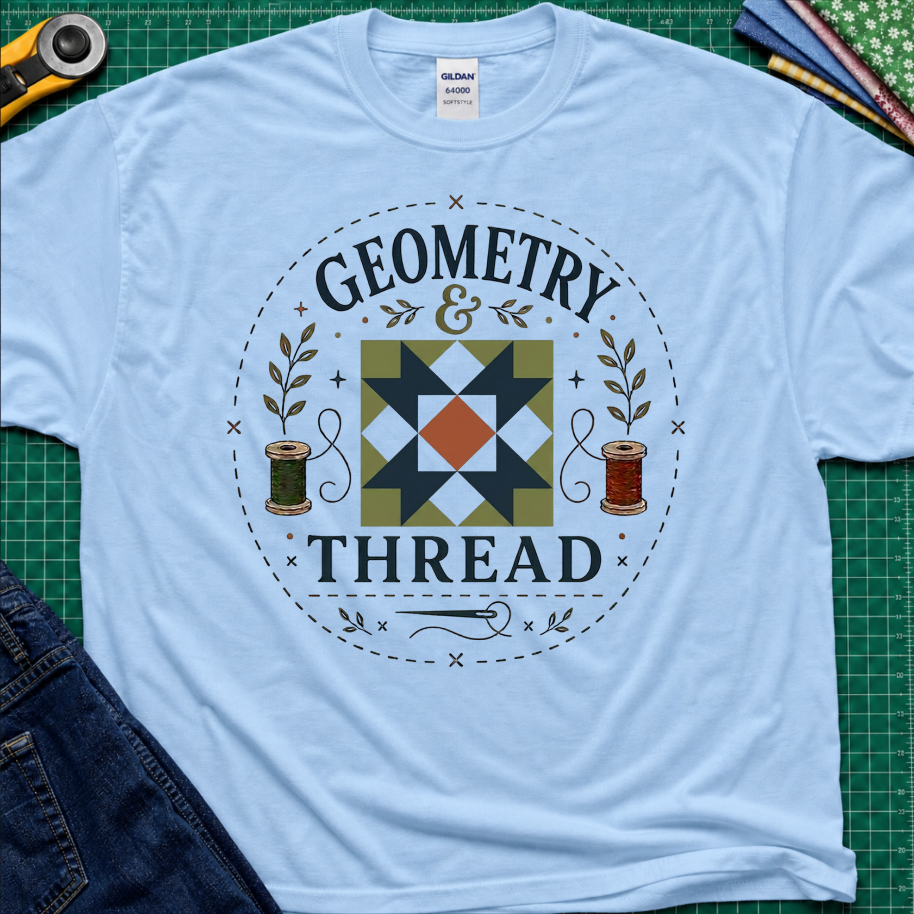 A light blue Gildan cotton t-shirt with a circular 'Geometry & Thread' graphic on the chest with quilt block, cotton spools and needle. Styled in a flat-lay on a green cutting mat with sewing supplies.