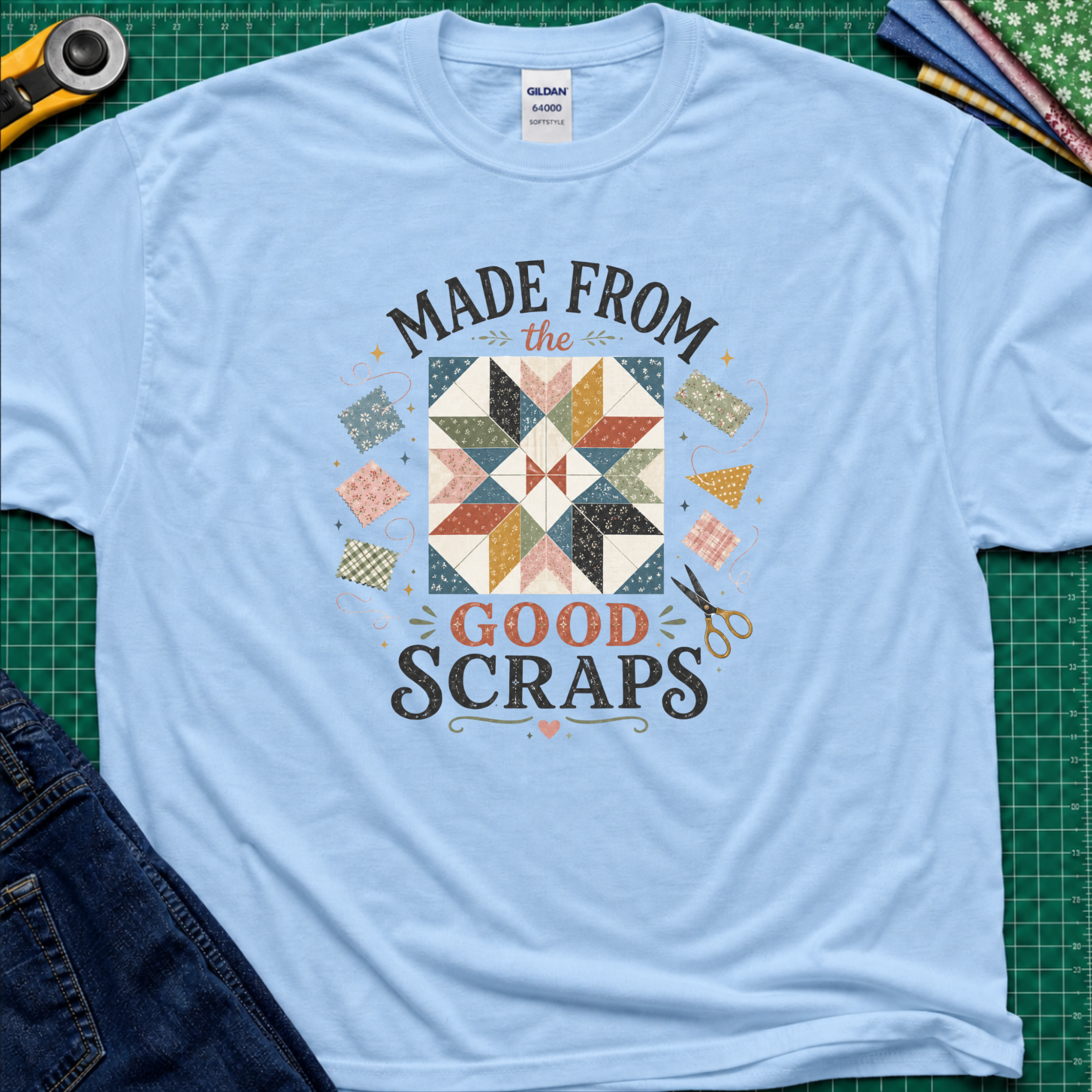 A light blue Gildan t-shirt with a retro-style quilted scrapbook graphic and the text 'MADE FROM the GOOD SCRAPS'. The shirt is styled in a flat-lay on a green cutting mat with sewing tools and fabric.