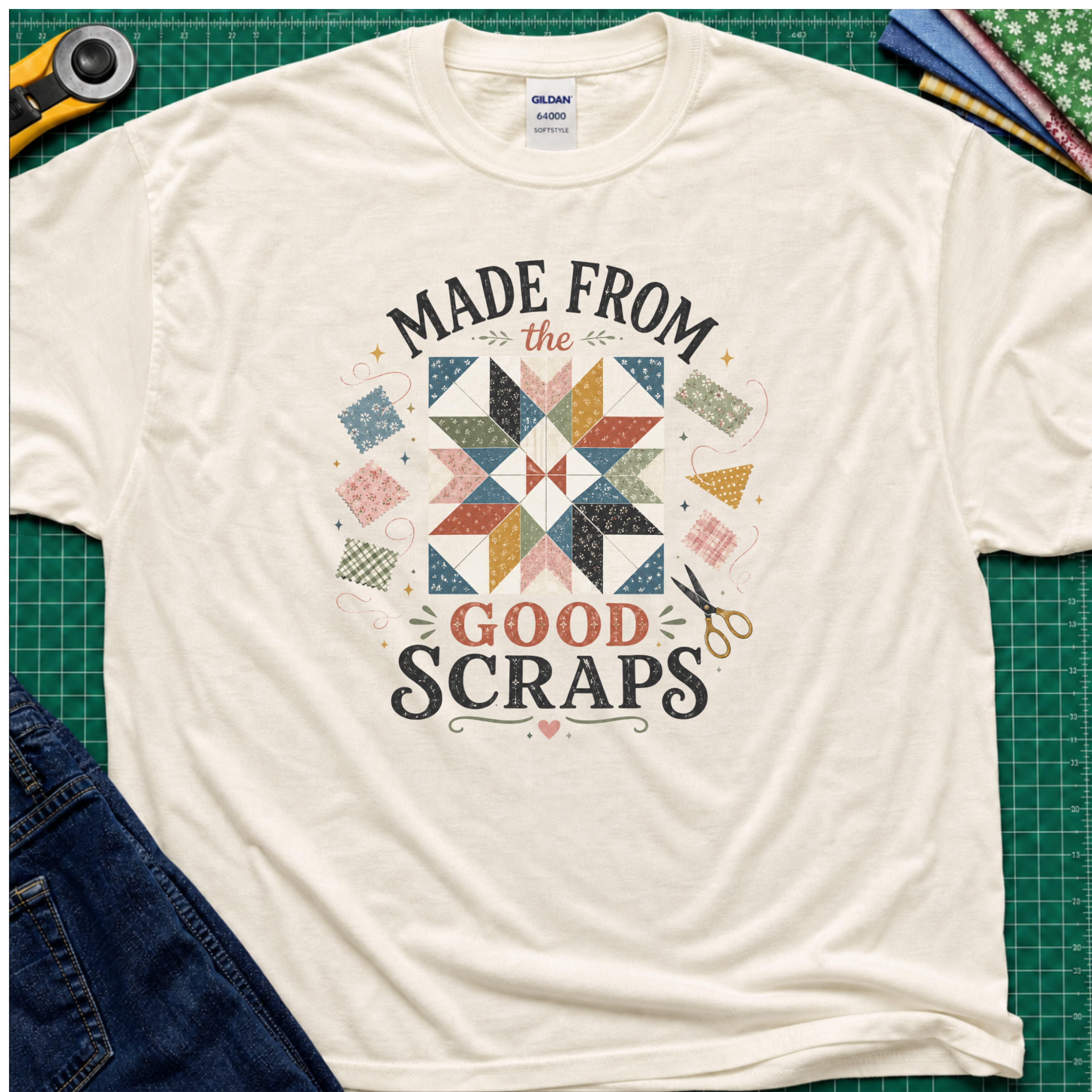 A light cream Gildan t-shirt with a retro-style quilted scrapbook graphic and the text 'MADE FROM the GOOD SCRAPS'. The shirt is styled in a flat-lay on a green cutting mat with sewing tools and fabric.