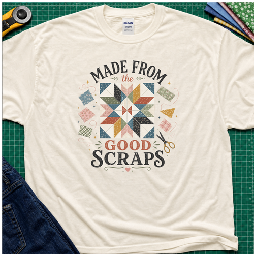 A light cream Gildan t-shirt with a retro-style quilted scrapbook graphic and the text 'MADE FROM the GOOD SCRAPS'. The shirt is styled in a flat-lay on a green cutting mat with sewing tools and fabric.
