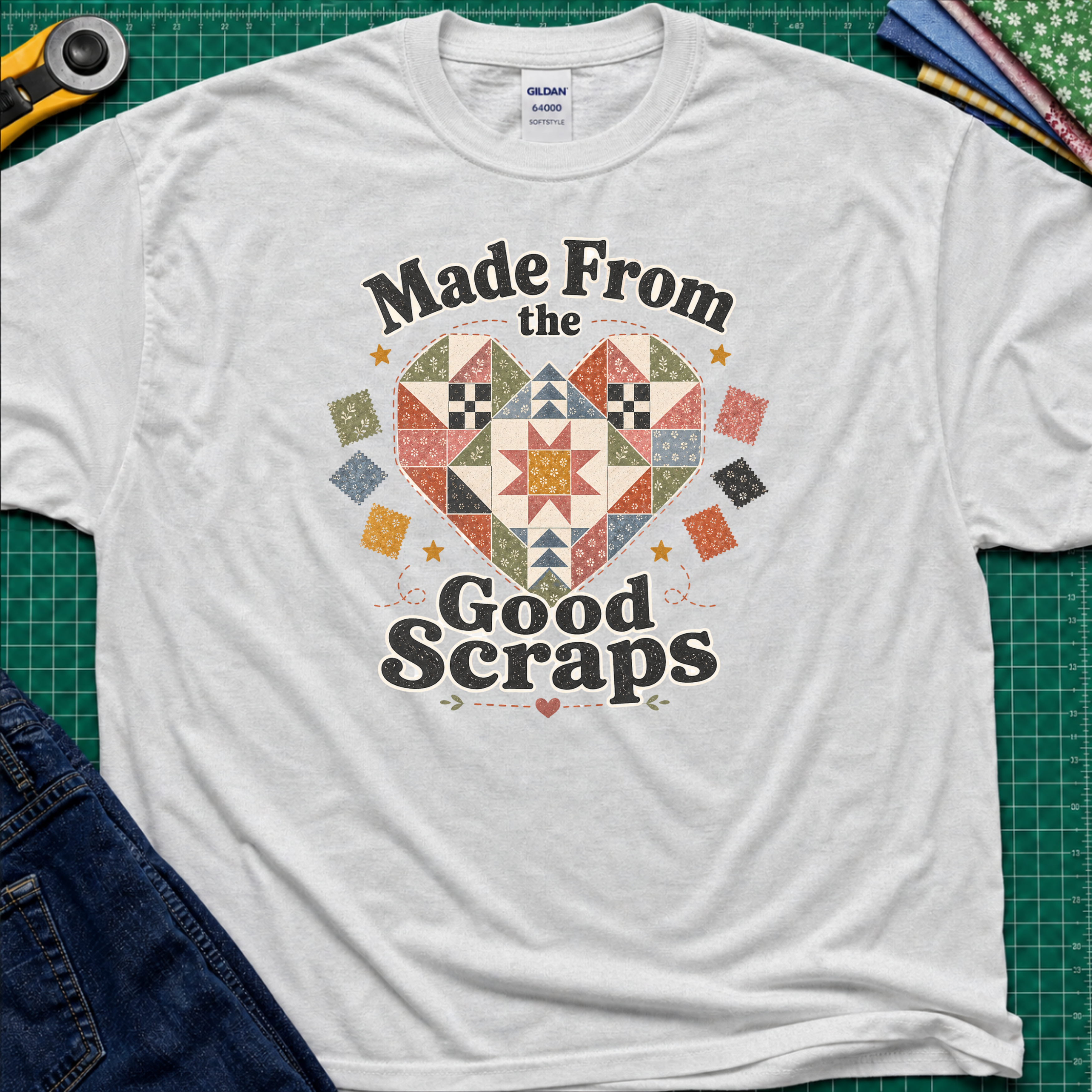 An ice grey Gildan t-shirt with a colorful quilted heart graphic and the text 'Made From the Good Scraps' printed on the front, styled flat on a green cutting mat with sewing tools and denim jeans.