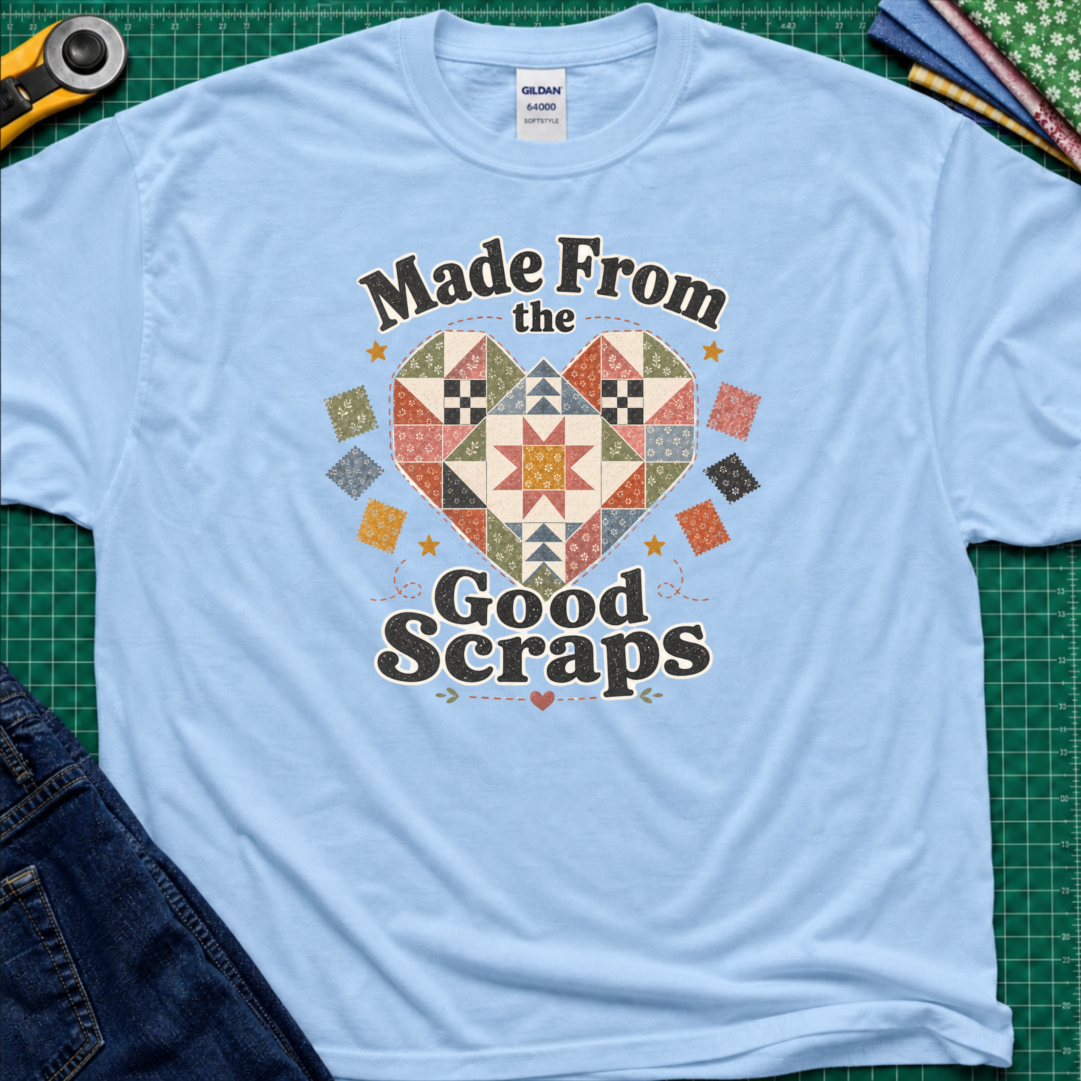 A light blue Gildan t-shirt with a colorful quilted heart graphic and the text 'Made From the Good Scraps' printed on the front, styled flat on a green cutting mat with sewing tools and denim jeans.