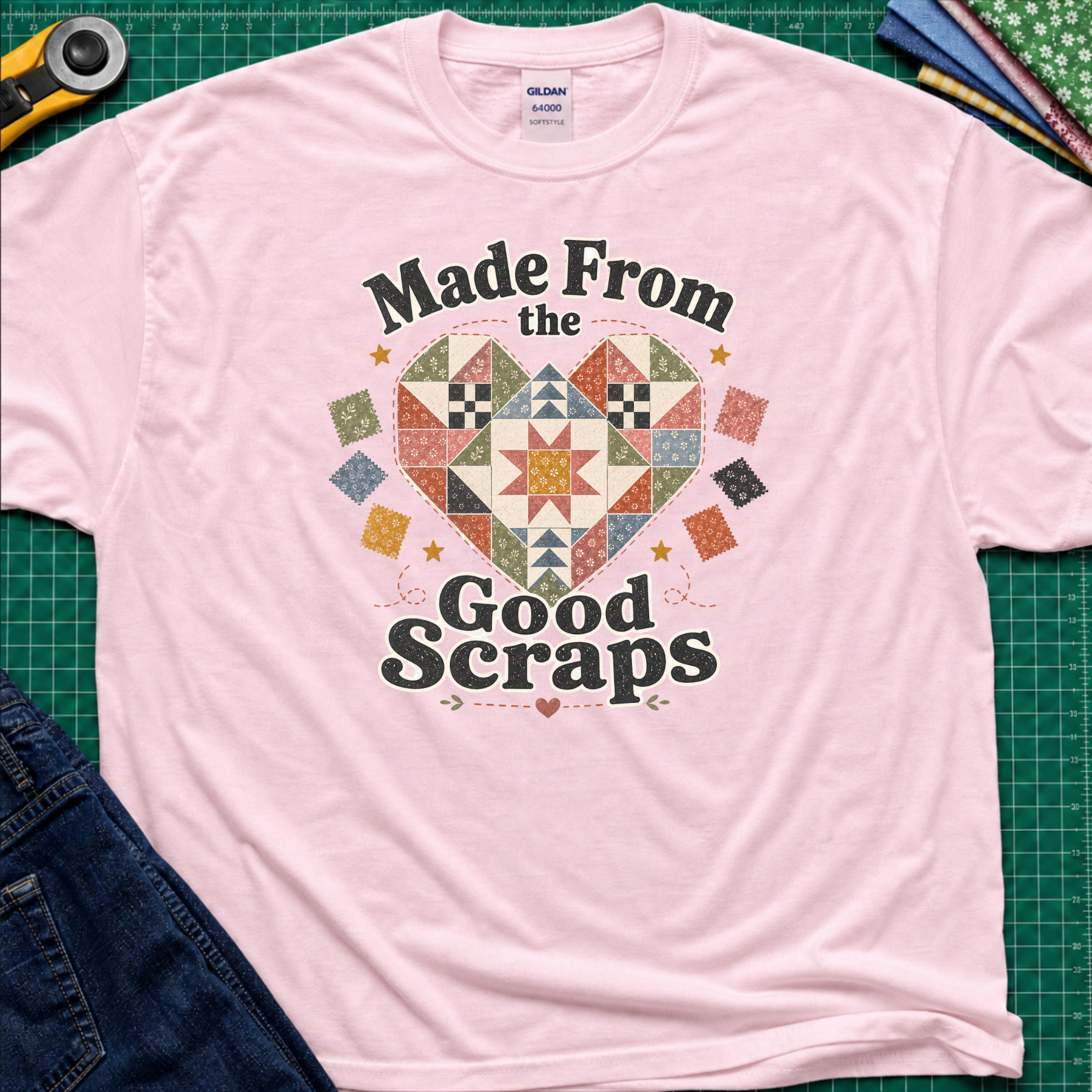 A light pink Gildan t-shirt with a colorful quilted heart graphic and the text 'Made From the Good Scraps' printed on the front, styled flat on a green cutting mat with sewing tools and denim jeans.
