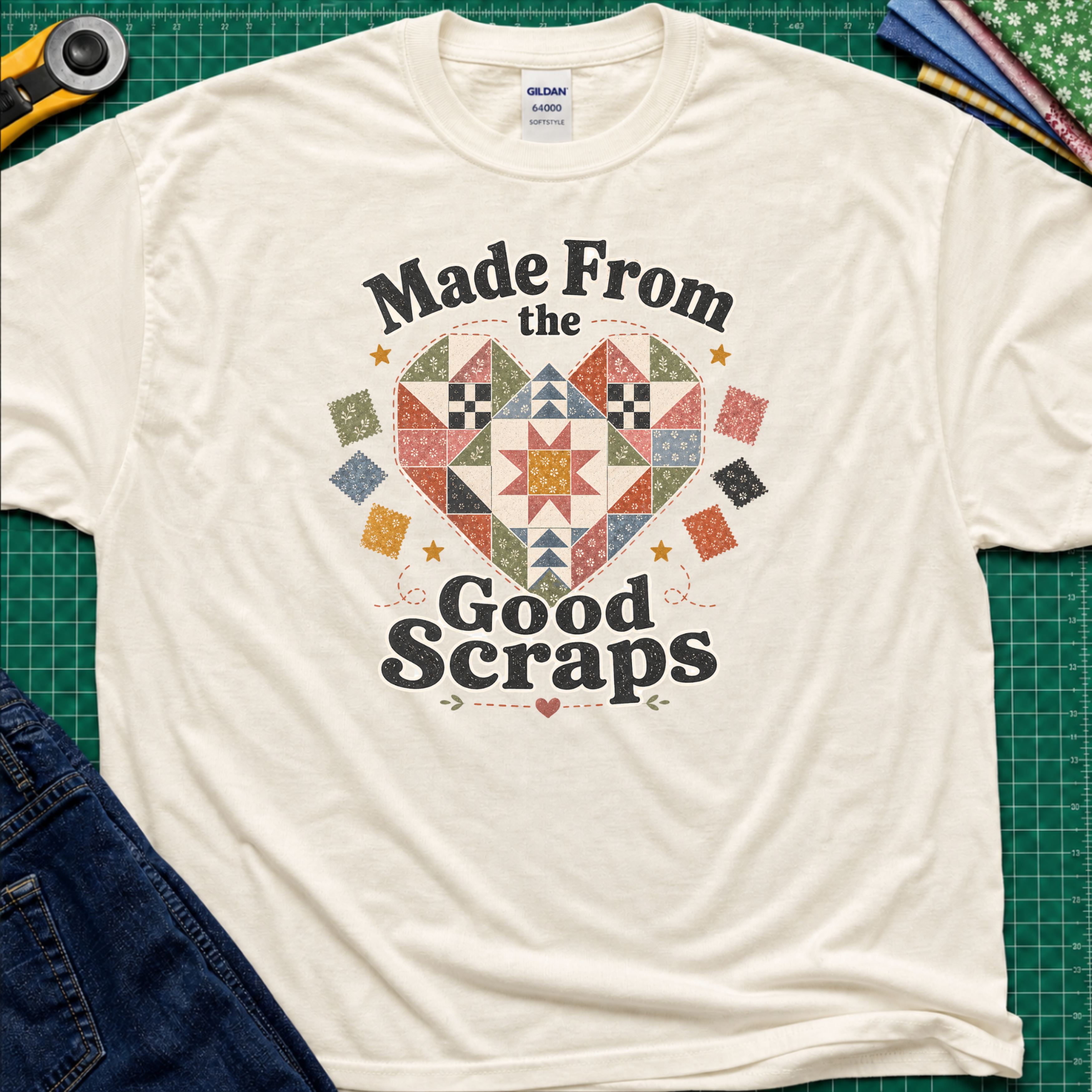 A light cream Gildan t-shirt with a colorful quilted heart graphic and the text 'Made From the Good Scraps' printed on the front, styled flat on a green cutting mat with sewing tools and denim jeans.