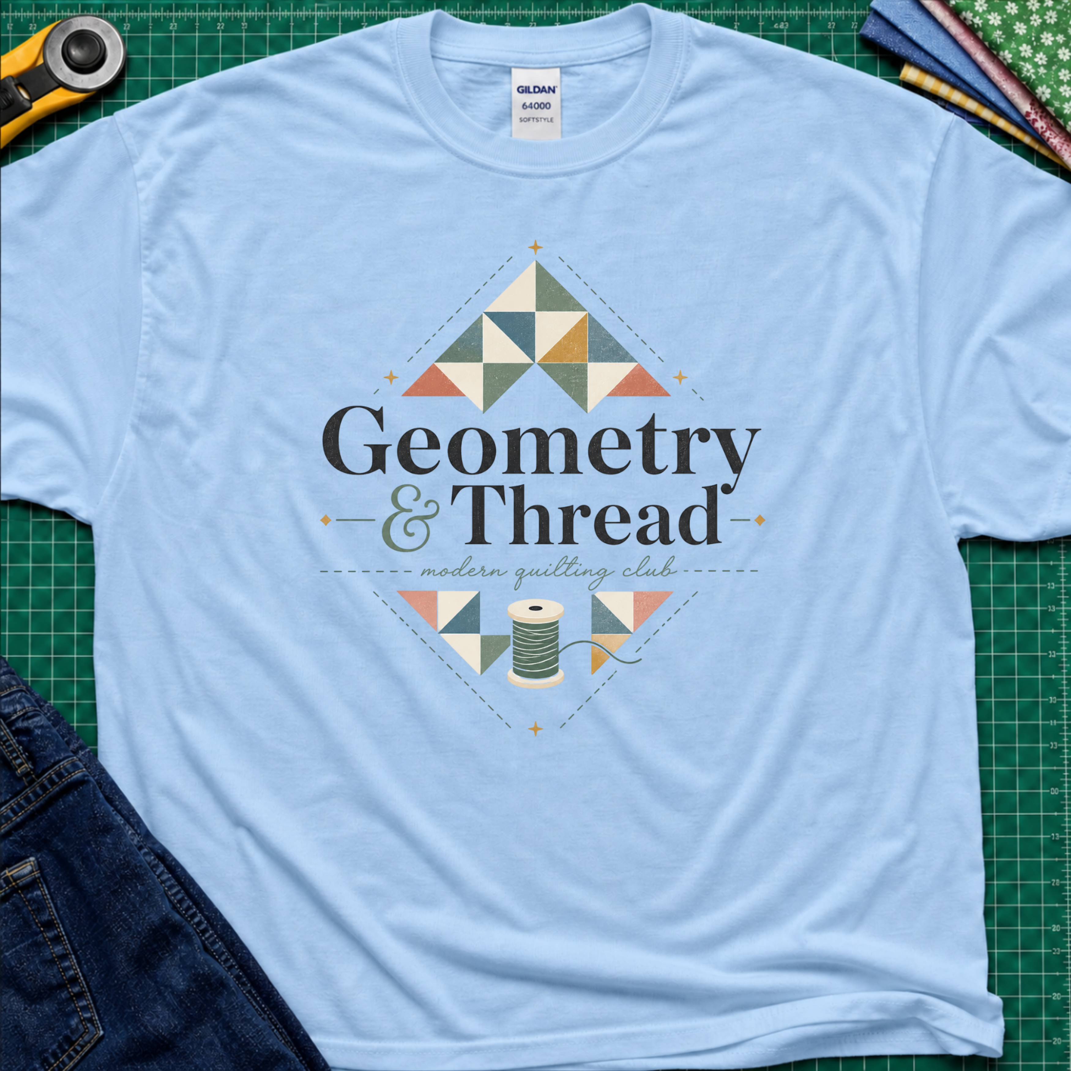 A light blue Gildan t-shirt with a colorful geometric quilt pattern and the text 'Geometry & Thread - modern quilting club' printed on the front. The shirt is styled in a flat-lay on a green cutting mat with sewing props like a rotary cutter, fabrics, and jeans.