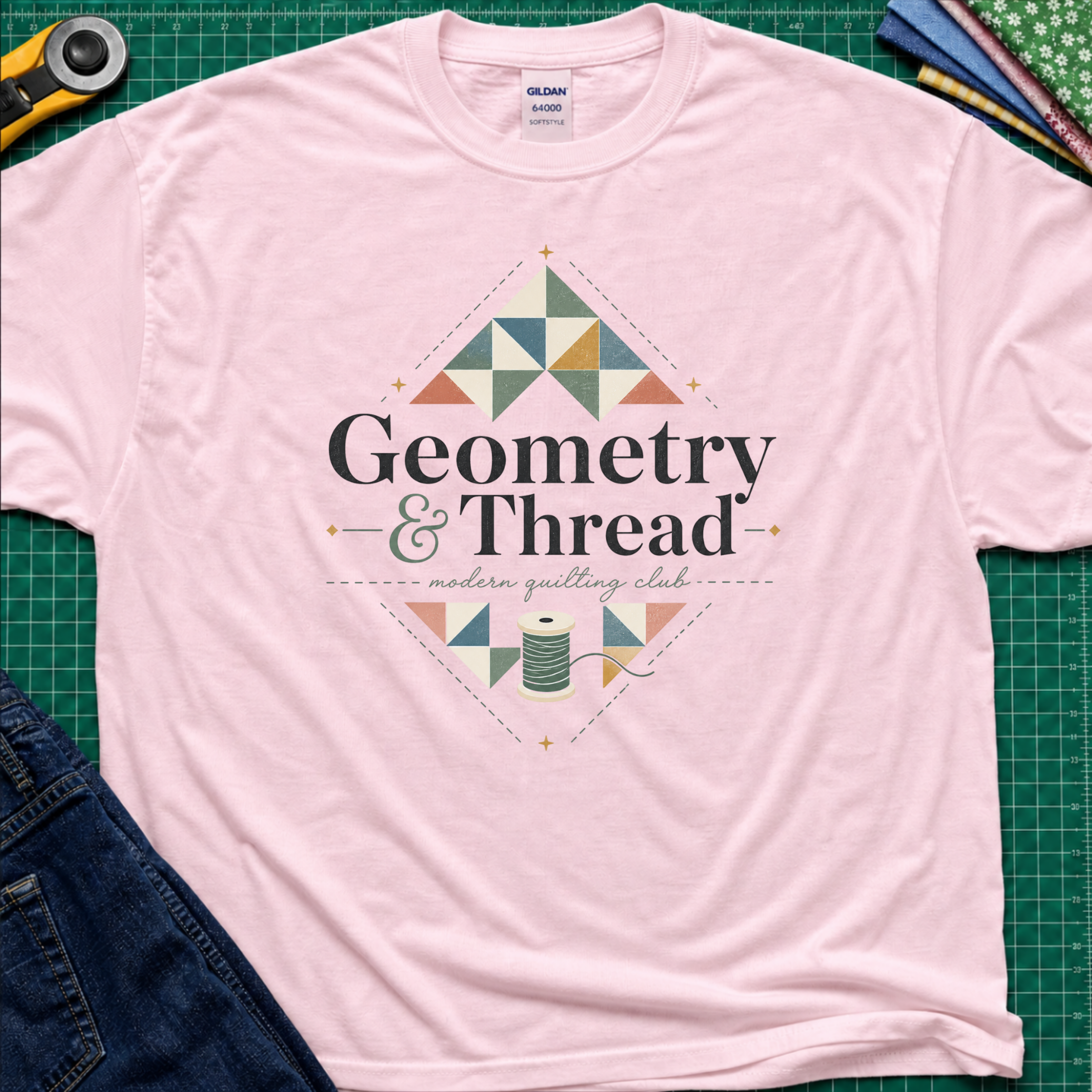 A light pink Gildan t-shirt with a colorful geometric quilt pattern and the text 'Geometry & Thread - modern quilting club' printed on the front. The shirt is styled in a flat-lay on a green cutting mat with sewing props like a rotary cutter, fabrics, and jeans.