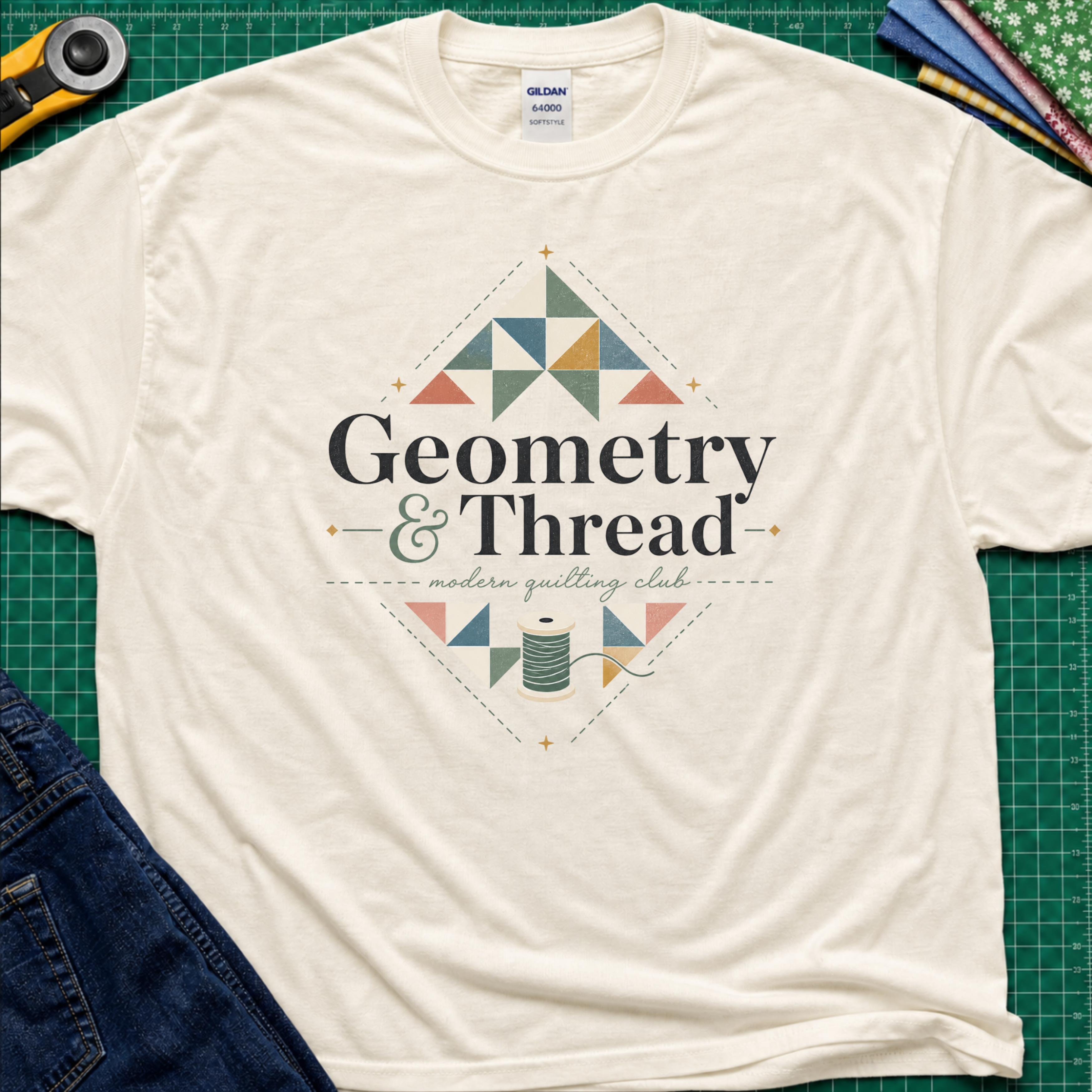 A light cream Gildan t-shirt with a colorful geometric quilt pattern and the text 'Geometry & Thread - modern quilting club' printed on the front. The shirt is styled in a flat-lay on a green cutting mat with sewing props like a rotary cutter, fabrics, and jeans.