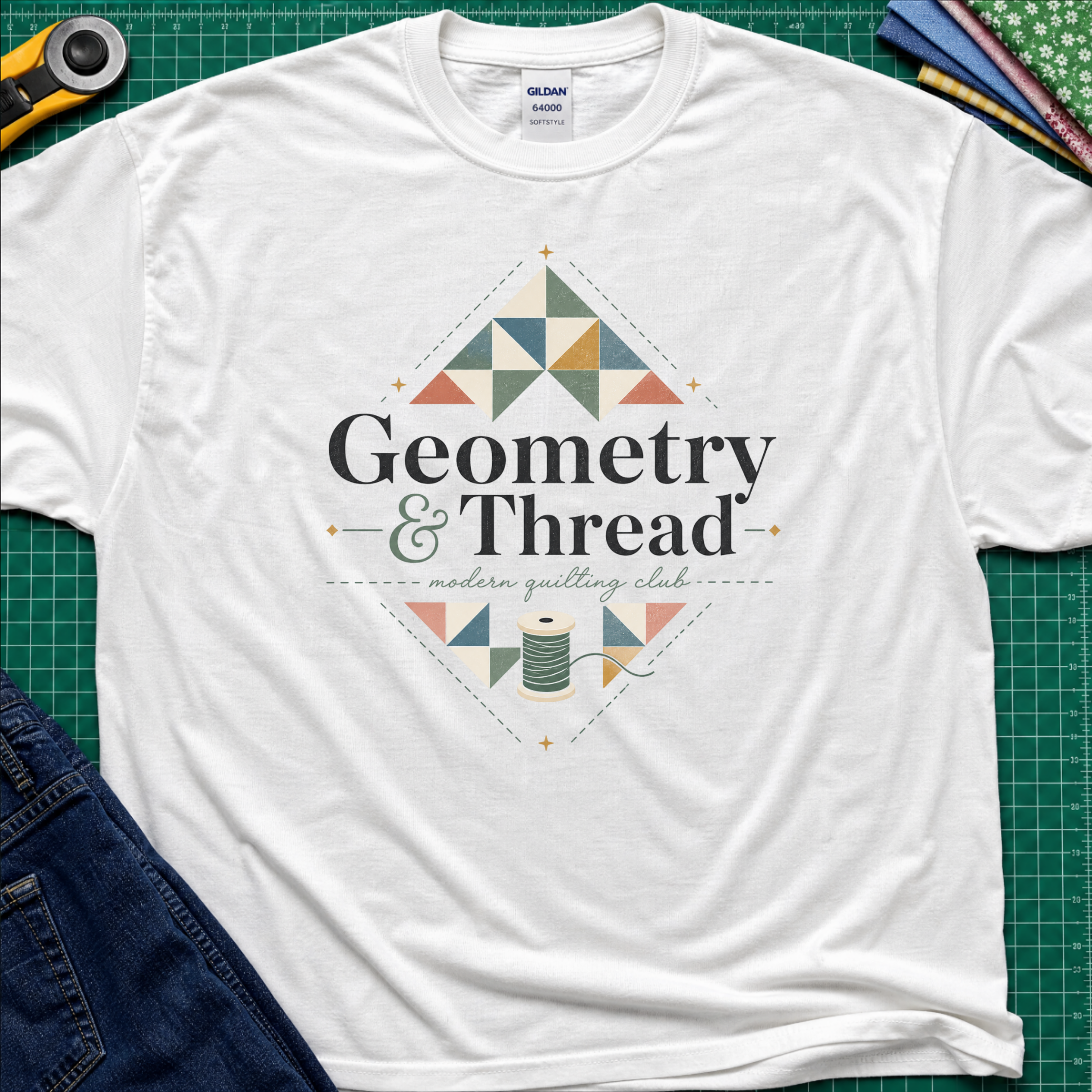 A white Gildan t-shirt with a colorful geometric quilt pattern and the text 'Geometry & Thread - modern quilting club' printed on the front. The shirt is styled in a flat-lay on a green cutting mat with sewing props like a rotary cutter, fabrics, and jeans.