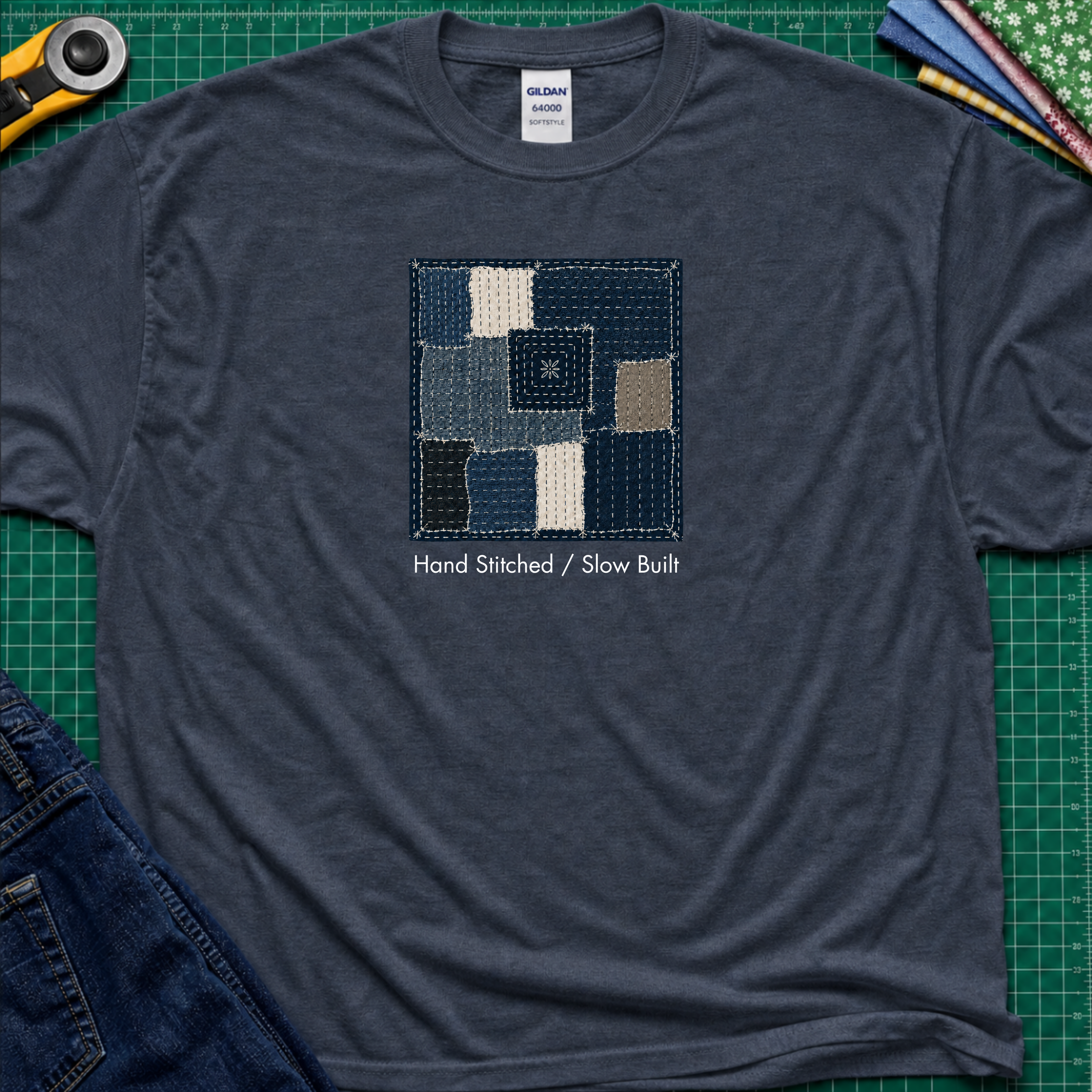 A heather navy blue Gildan t-shirt featuring a central graphic of a sashiko block made from  fabric squares in various shades of blue, navy, and beige, with the text 'Hand Stitched / Slow Built' underneath. The t-shirt lays flat on a green cutting mat with a rotary cutter, fabric stack and blue jeans partially visible in the corners.