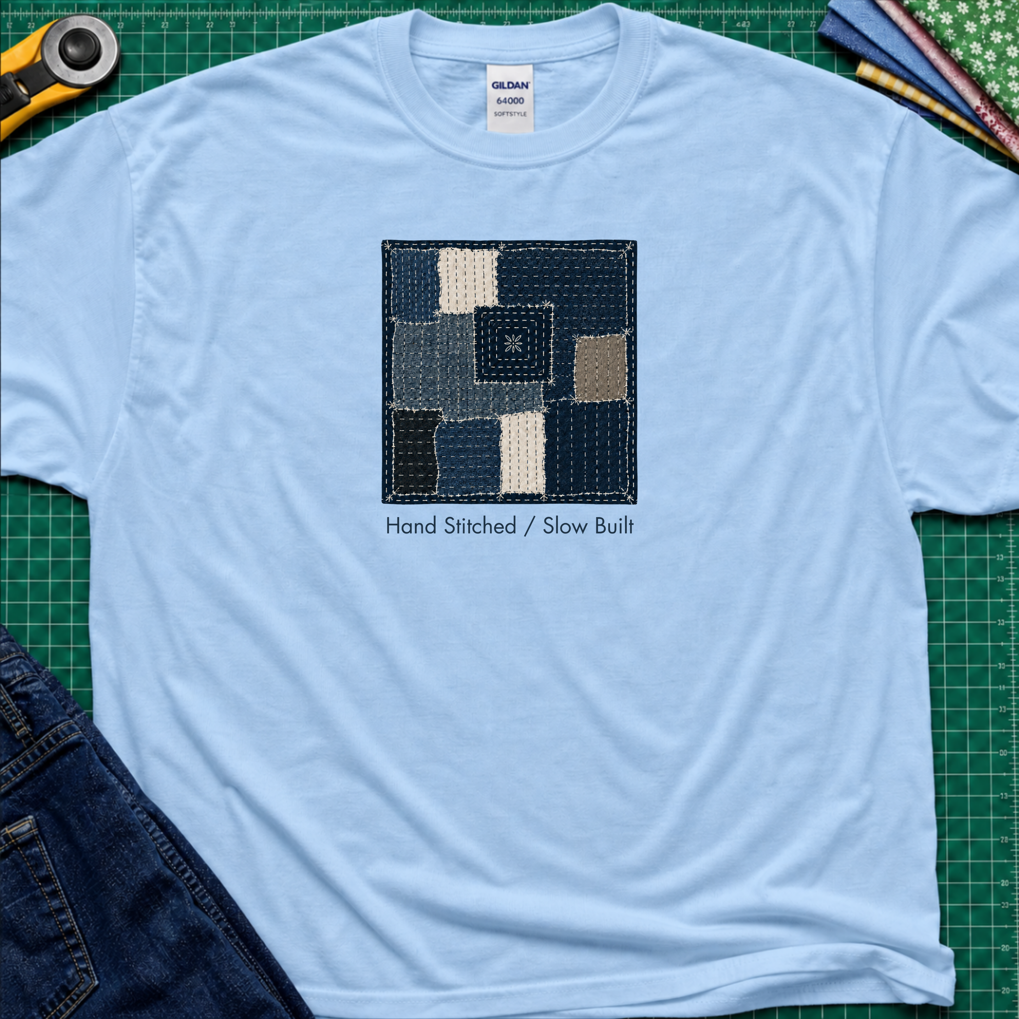 A light blue Gildan t-shirt featuring a central graphic of a sashiko block made from  fabric squares in various shades of blue, navy, and beige, with the text 'Hand Stitched / Slow Built' underneath. The t-shirt lays flat on a green cutting mat with a rotary cutter, fabric stack and blue jeans partially visible in the corners.