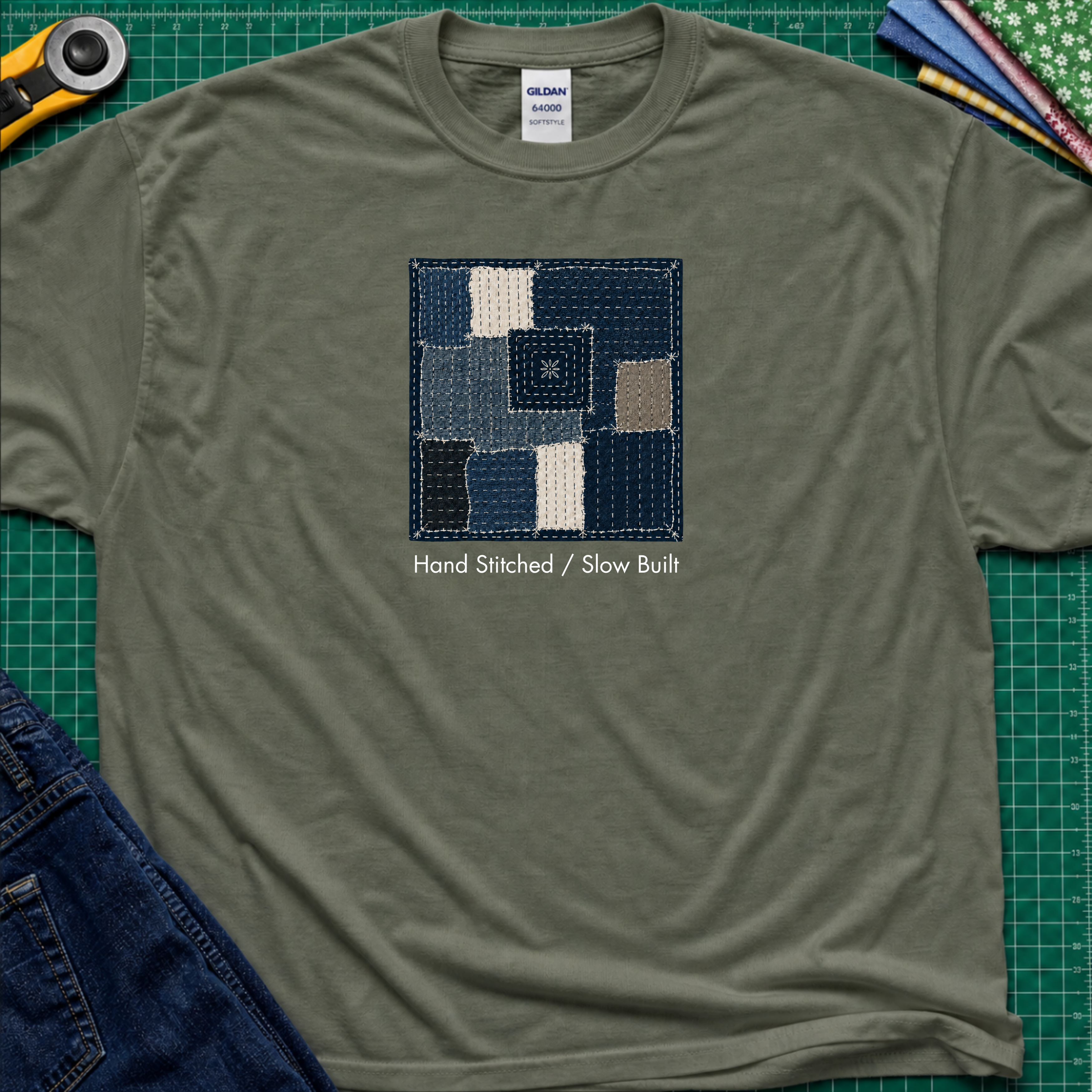 A military green Gildan t-shirt featuring a central graphic of a sashiko block made from  fabric squares in various shades of blue, navy, and beige, with the text 'Hand Stitched / Slow Built' underneath. The t-shirt lays flat on a green cutting mat with a rotary cutter, fabric stack and blue jeans partially visible in the corners.