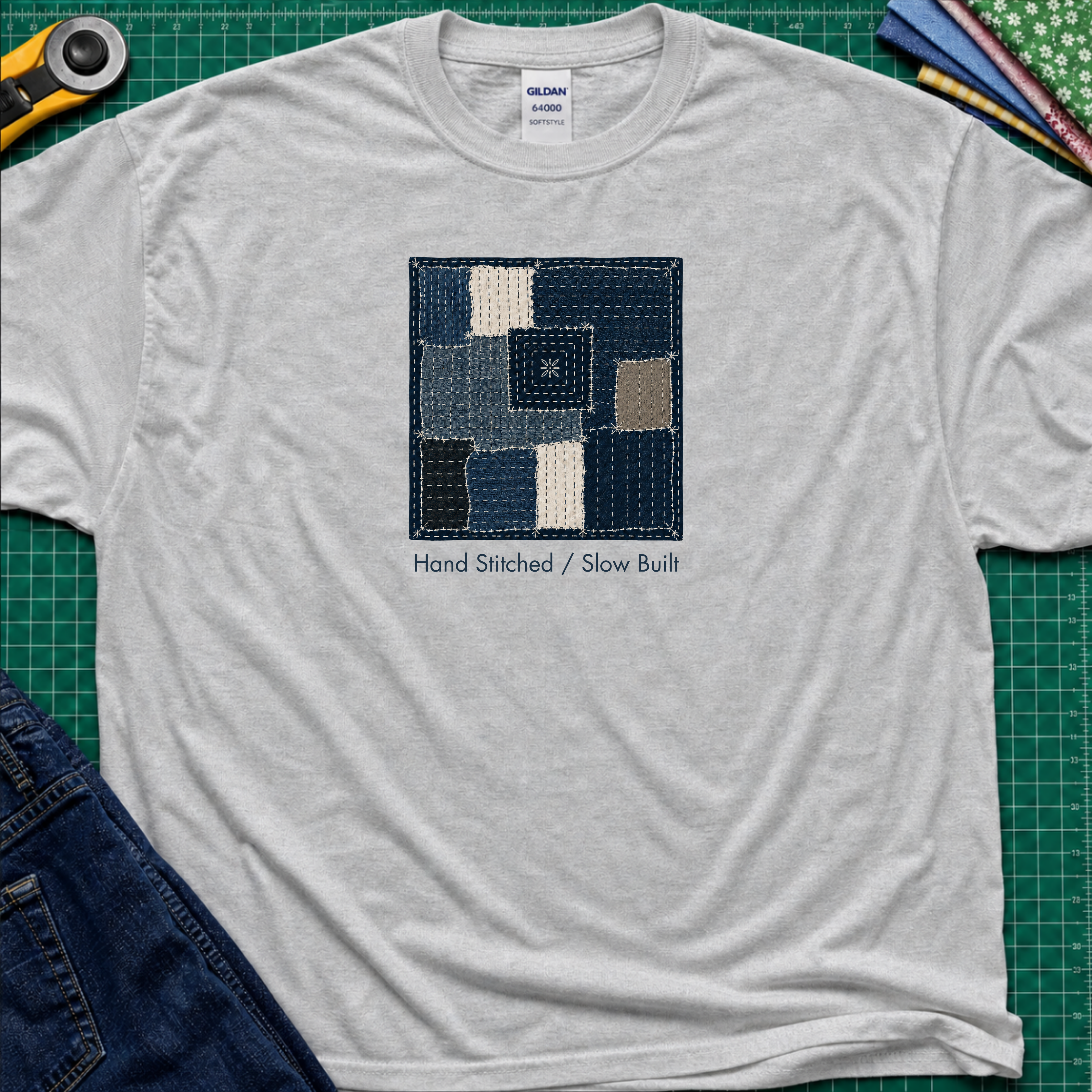 A sports grey Gildan t-shirt featuring a central graphic of a sashiko block made from  fabric squares in various shades of blue, navy, and beige, with the text 'Hand Stitched / Slow Built' underneath. The t-shirt lays flat on a green cutting mat with a rotary cutter, fabric stack and blue jeans partially visible in the corners.