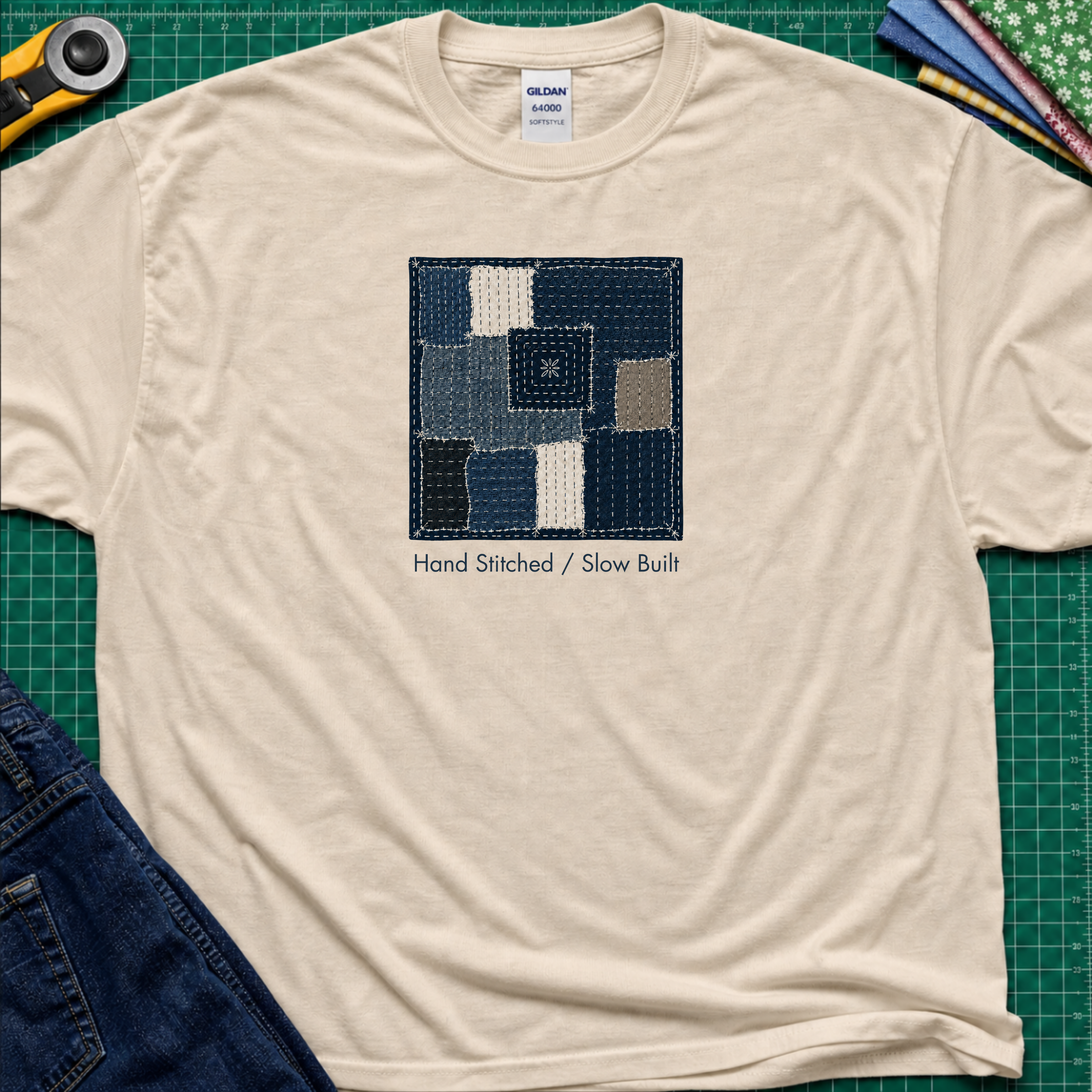 A dark cream Gildan t-shirt featuring a central graphic of a sashiko block made from  fabric squares in various shades of blue, navy, and beige, with the text 'Hand Stitched / Slow Built' underneath. The t-shirt lays flat on a green cutting mat with a rotary cutter, fabric stack and blue jeans partially visible in the corners.