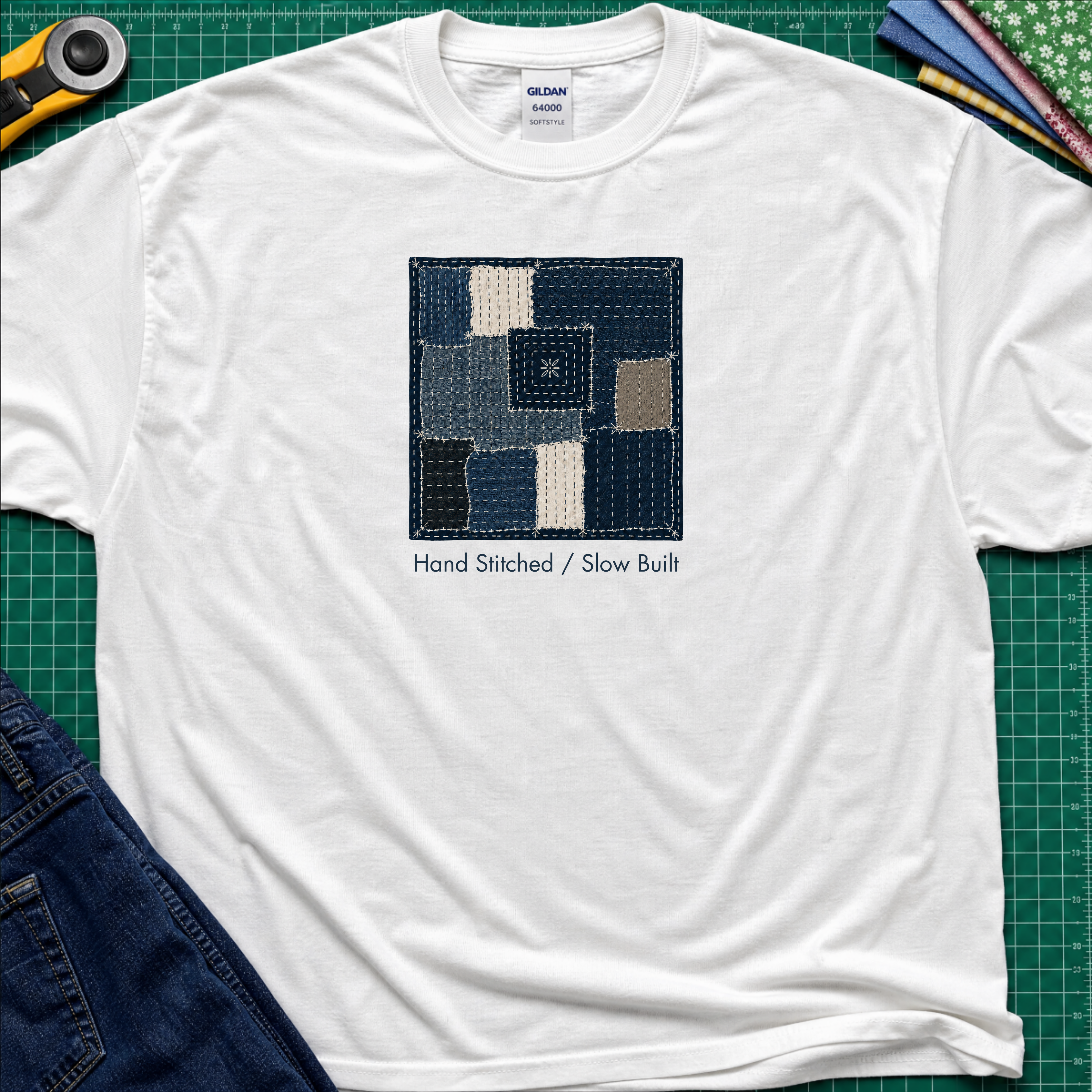 A white Gildan t-shirt featuring a central graphic of a sashiko block made from  fabric squares in various shades of blue, navy, and beige, with the text 'Hand Stitched / Slow Built' underneath. The t-shirt lays flat on a green cutting mat with a rotary cutter, fabric stack and blue jeans partially visible in the corners.