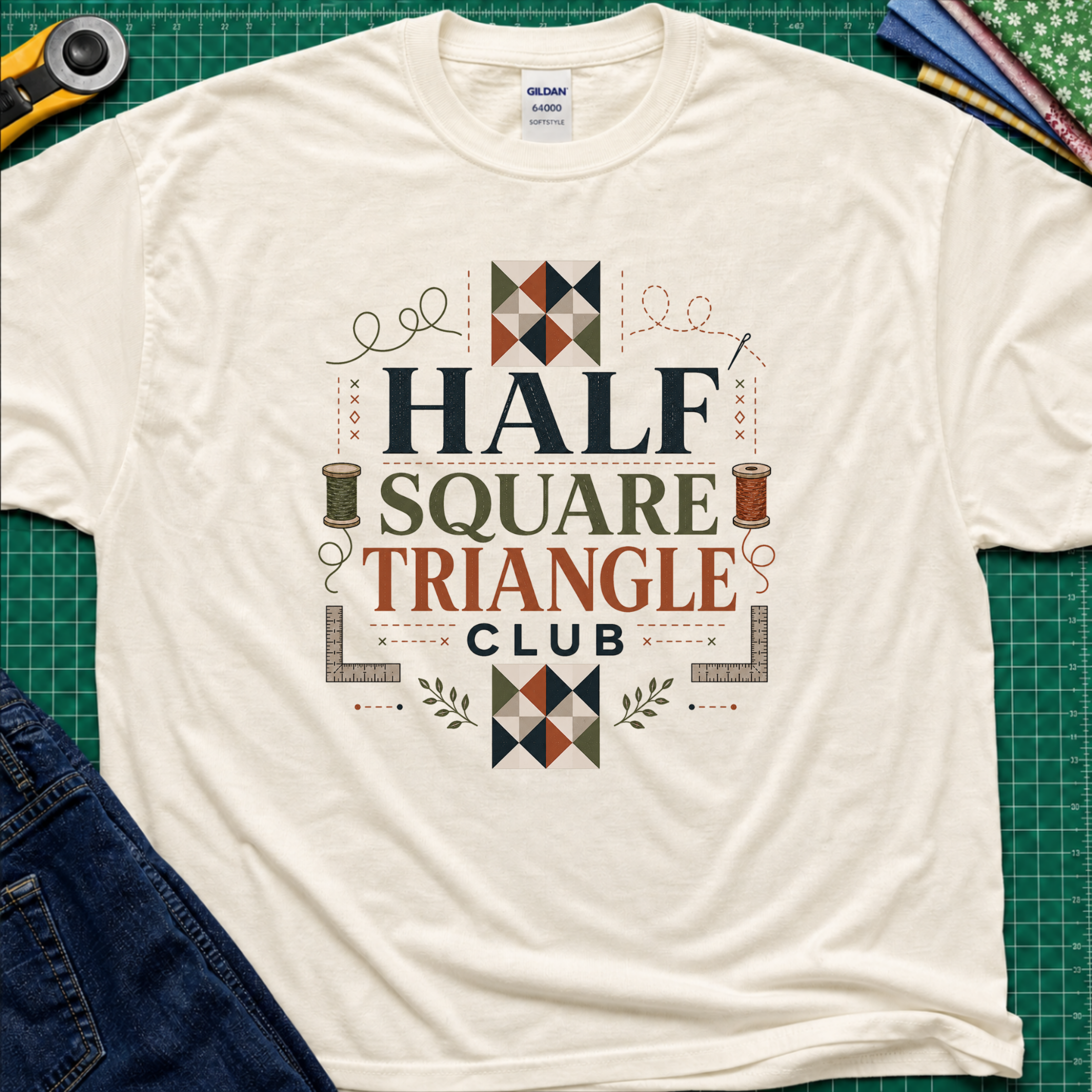 A light cream Gildan t-shirt with a 'Half Square Triangle Club' graphic, with quilt blocks, cotton spools and rulers. Laid flat on a green cutting mat with sewing props and jeans.