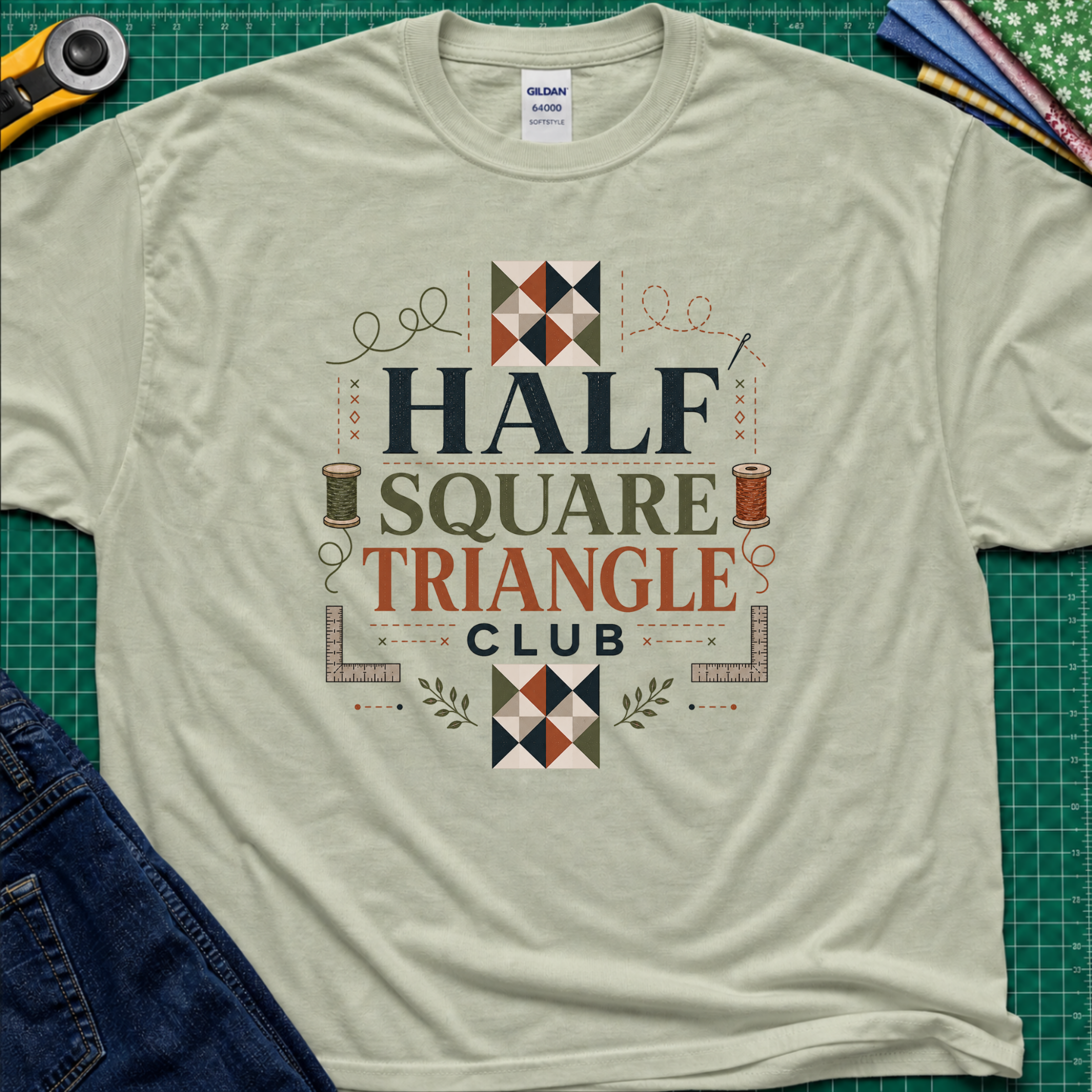 A light sage green Gildan t-shirt with a 'Half Square Triangle Club' graphic, with quilt blocks, cotton spools and rulers. Laid flat on a green cutting mat with sewing props and jeans.